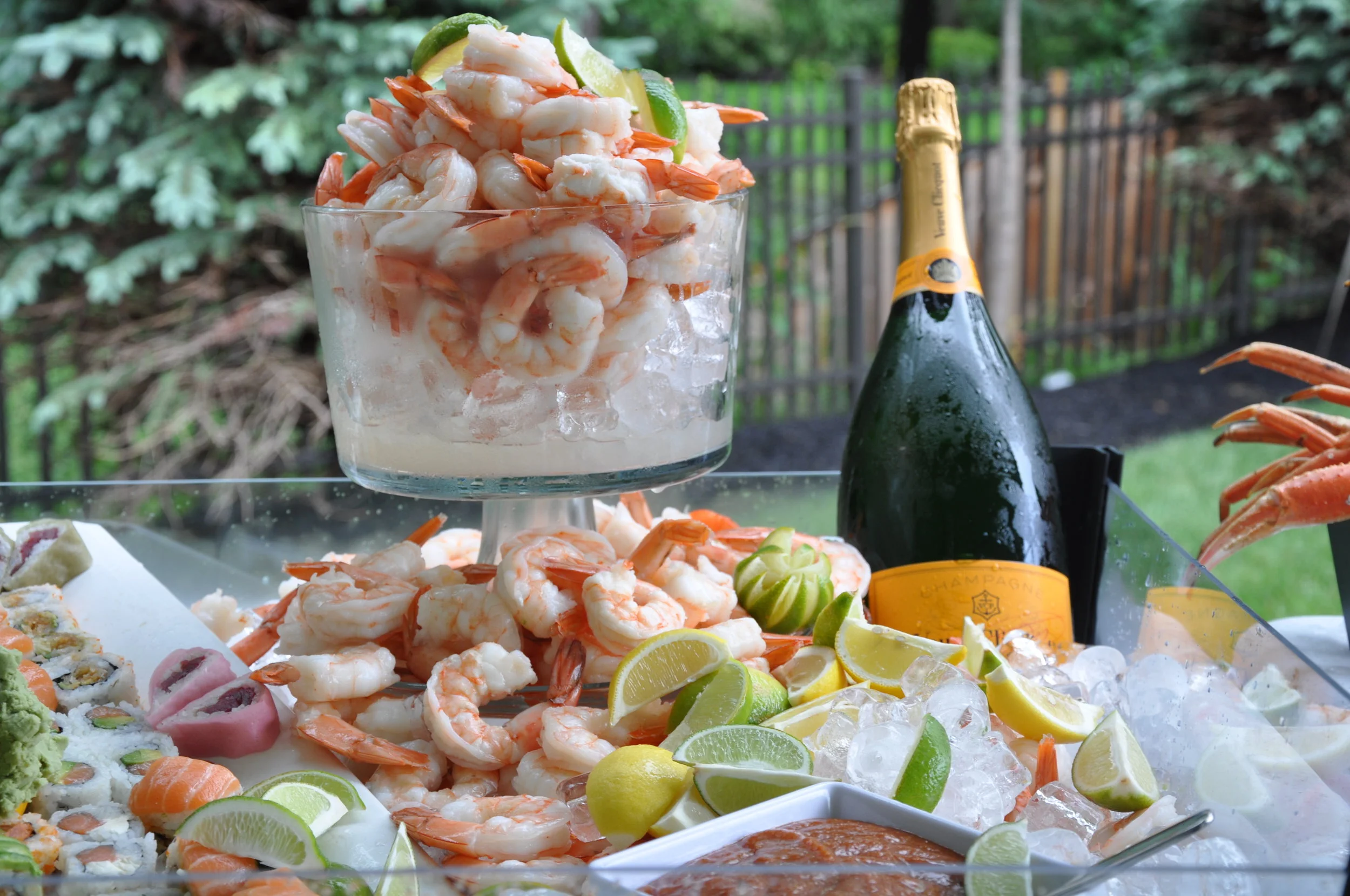 A glass bowl filled with cooked shrimp, garnished with lime wedges. Surrounding the bowl are more shrimp, sliced lemons, and various sushi rolls on ice, with a bottle of champagne nearby in an outdoor setting with trees and a wooden fence.