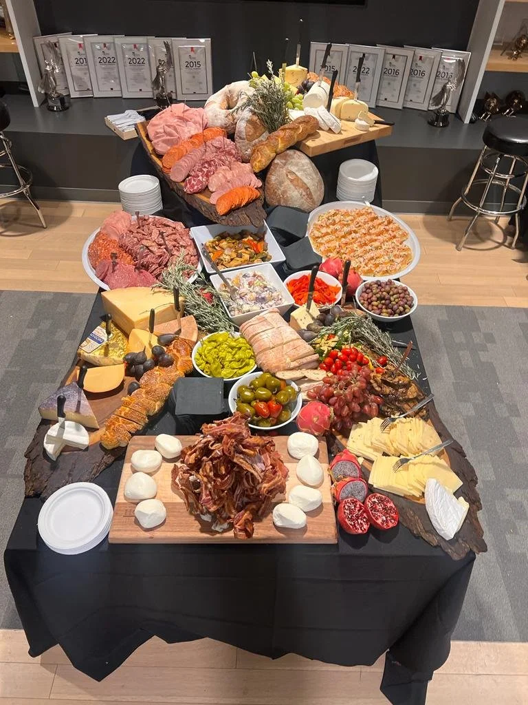Buffet table with assorted cheeses, cured meats, olives, pickles, bread, and fresh produce, set in a room with awards displayed on the wall.