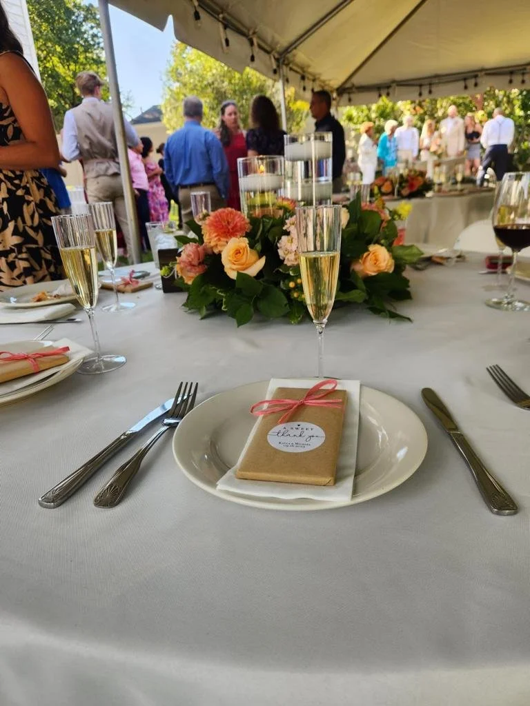 Elegant outdoor wedding reception table with floral centerpiece, champagne glasses, and wrapped favor with a pink bow, under a tent with guests in the background.