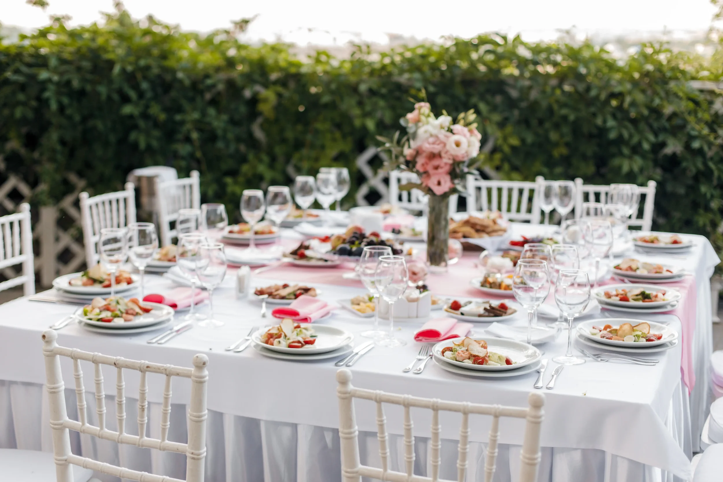 A long dining table set for a formal outdoor event, decorated with a pink tablecloth, white plates, glasses, silverware, and a pink floral centerpiece, with white chairs and greenery in the background.