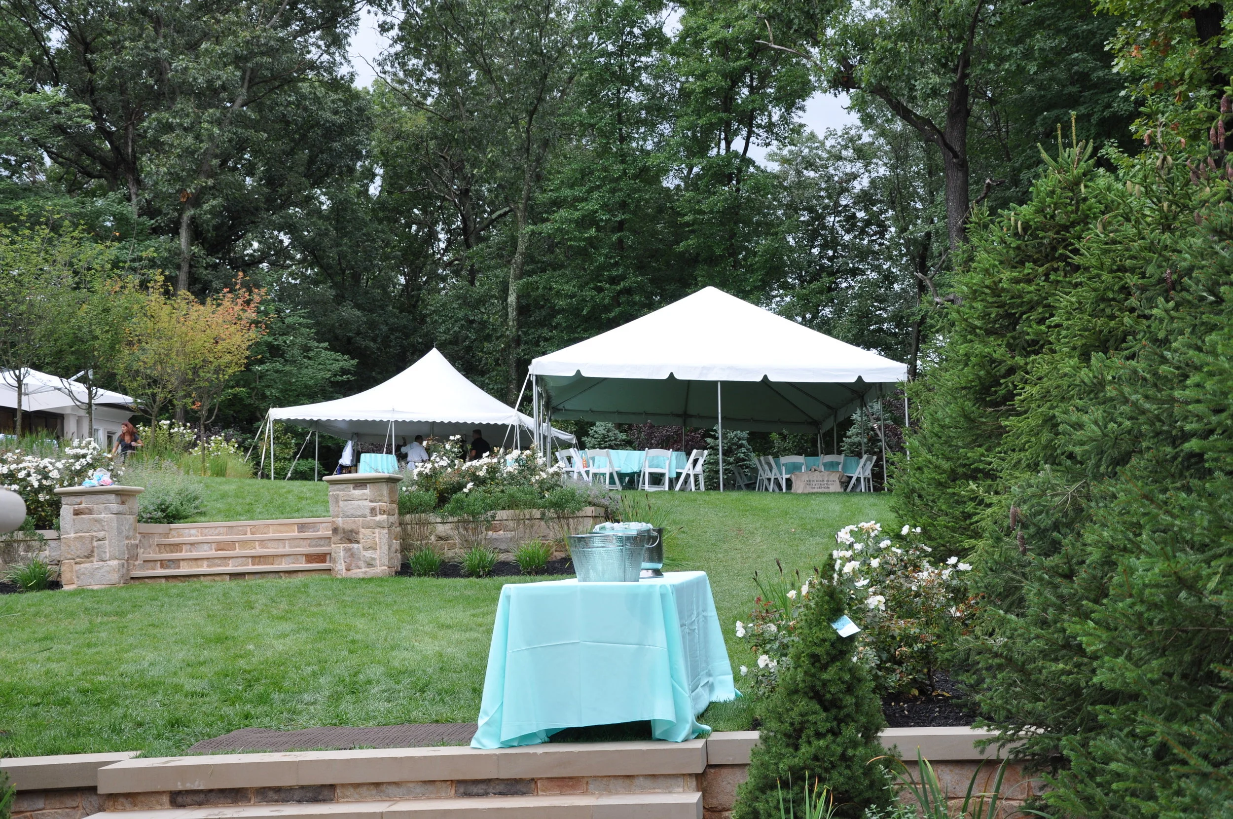 An outdoor garden set up with white tents, tables, and chairs for an event, surrounded by lush green trees and plants.