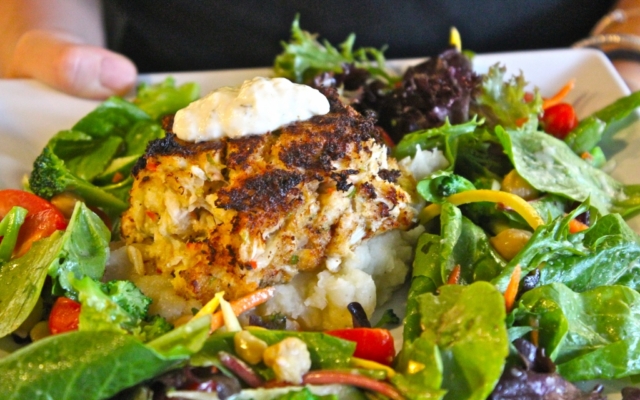 A plate with mashed potatoes topped with a crab cake and a dollop of tartar sauce, served with a side salad of mixed greens and vegetables.