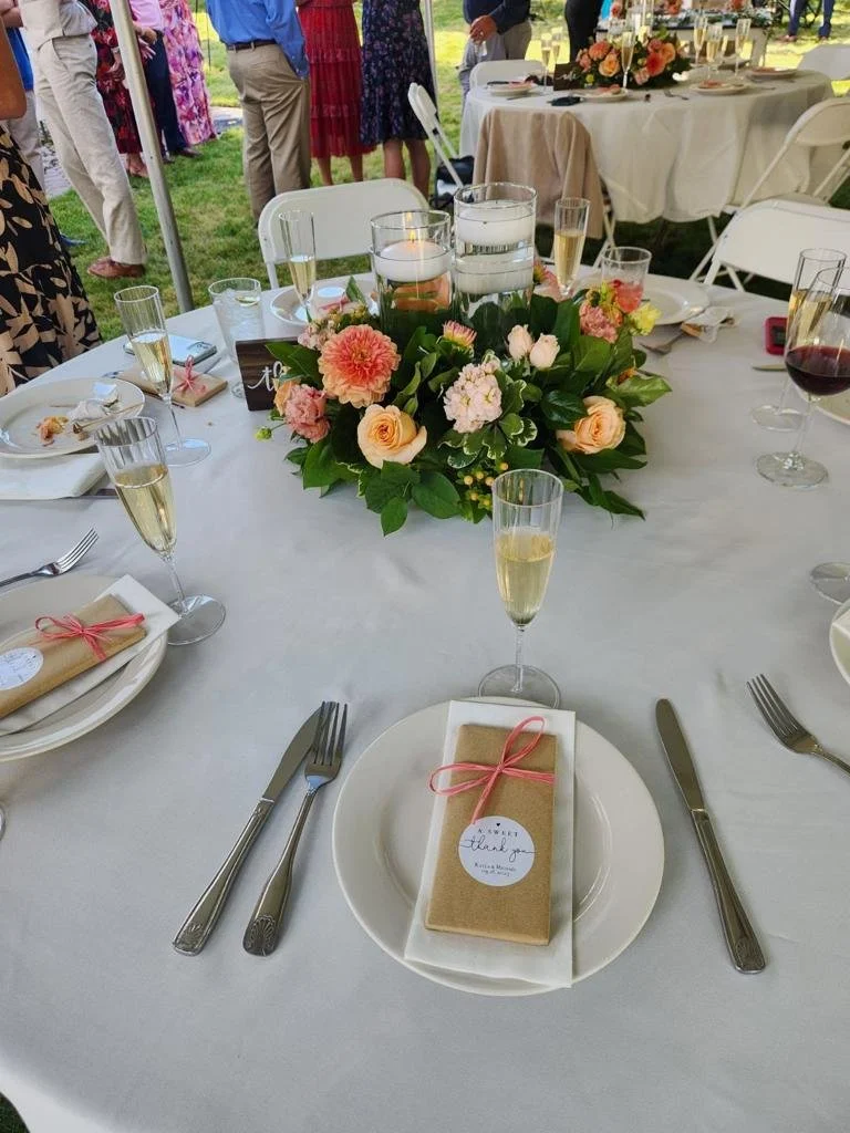 Table set for a celebration with floral centerpiece, champagne and wine glasses, wrapped favors, and a 'thank you' tag, outdoors with people in the background.