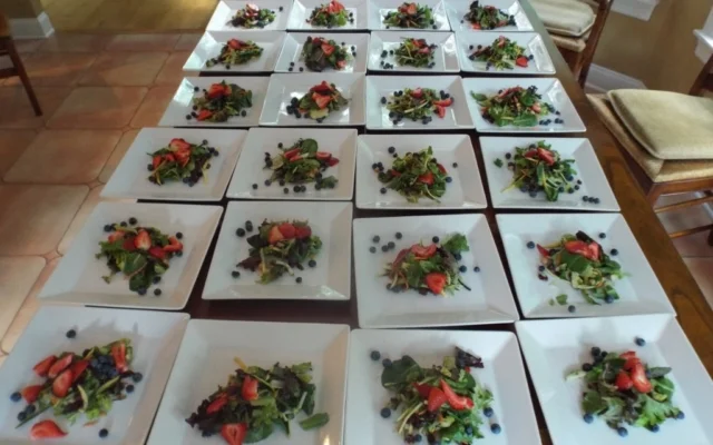 Multiple white plates with salads containing mixed greens, strawberries, and blueberries arranged on a wooden table.
