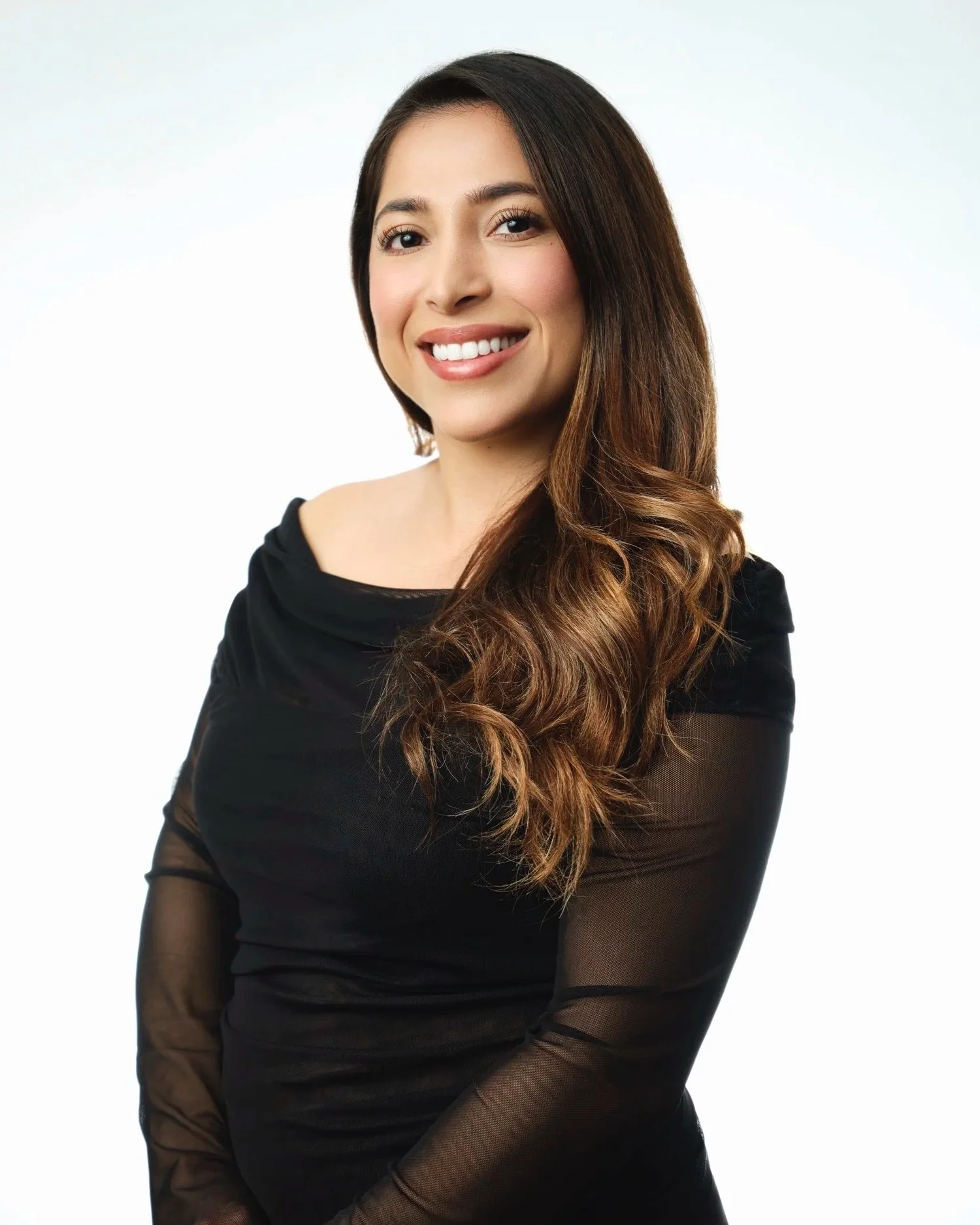 Portrait of a young woman with long wavy brown hair, smiling, wearing a black top with sheer sleeves, against a plain white background.