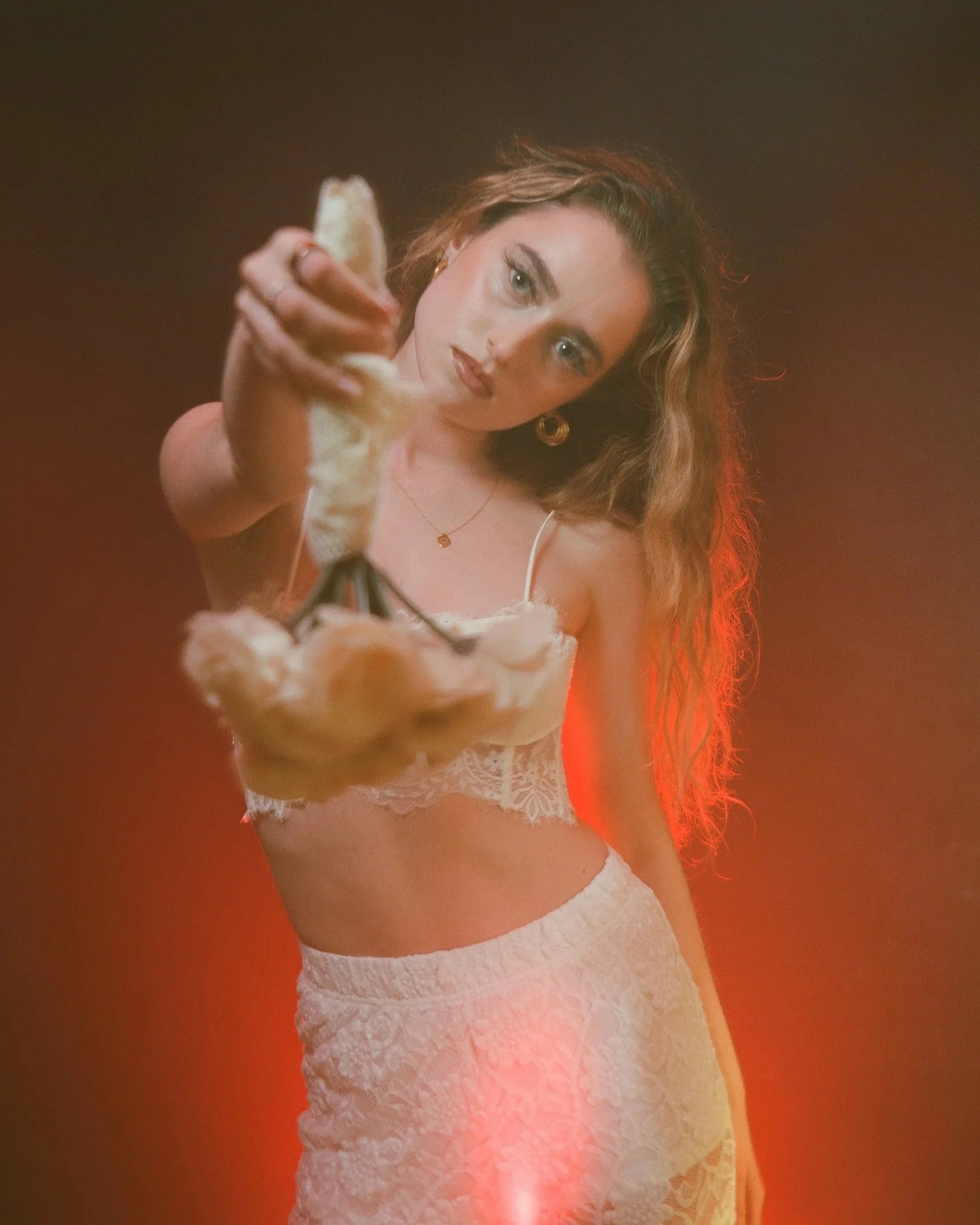 Young woman with long wavy hair, wearing a white lace bralette and matching skirt, holding a cheeseburger towards the camera with a serious expression, red lighting background.