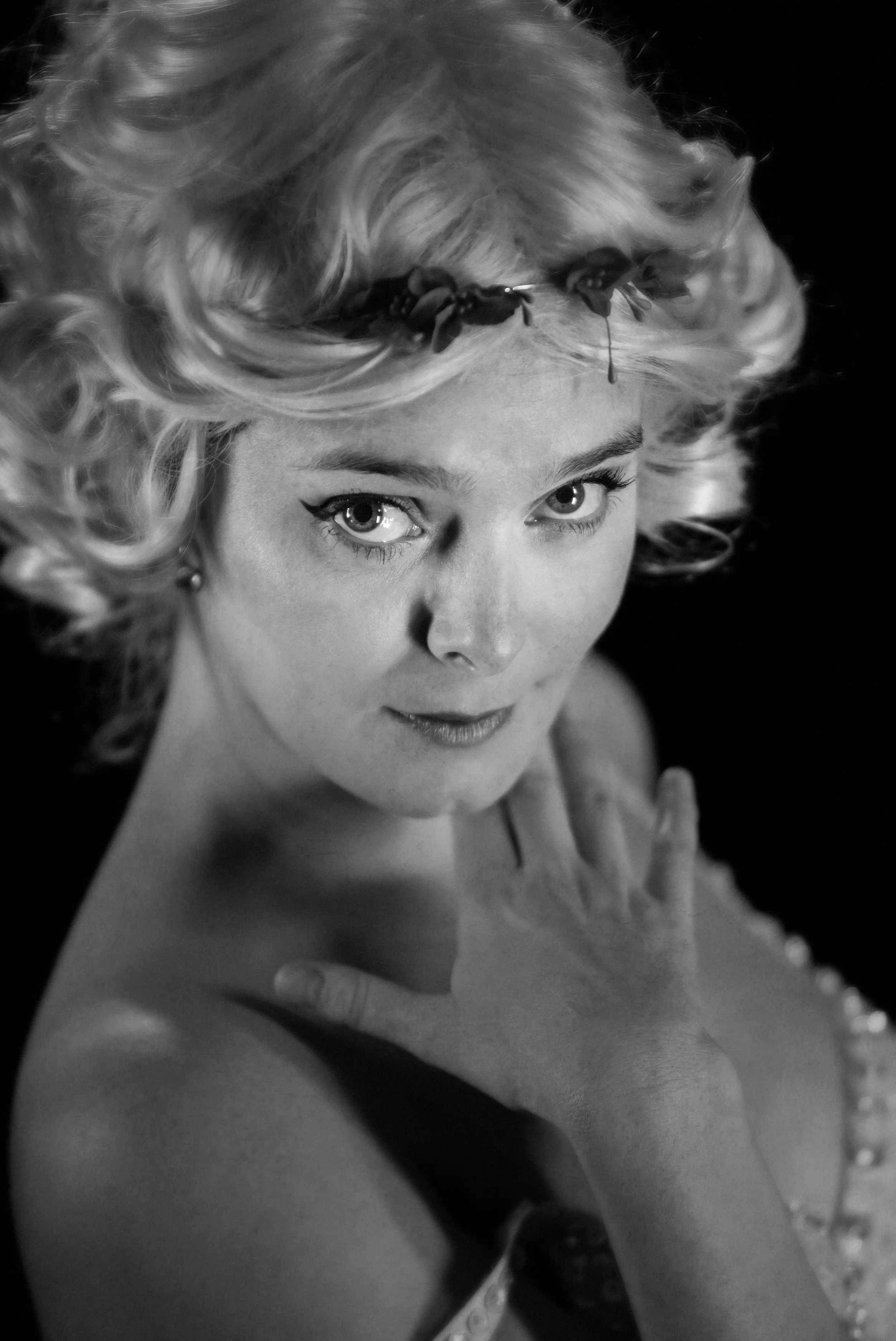 A black-and-white portrait of a woman with curly hair, looking directly at the camera, with her hand on her shoulder, Annoying in a thoughtful pose, with dark makeup and a confident expression.