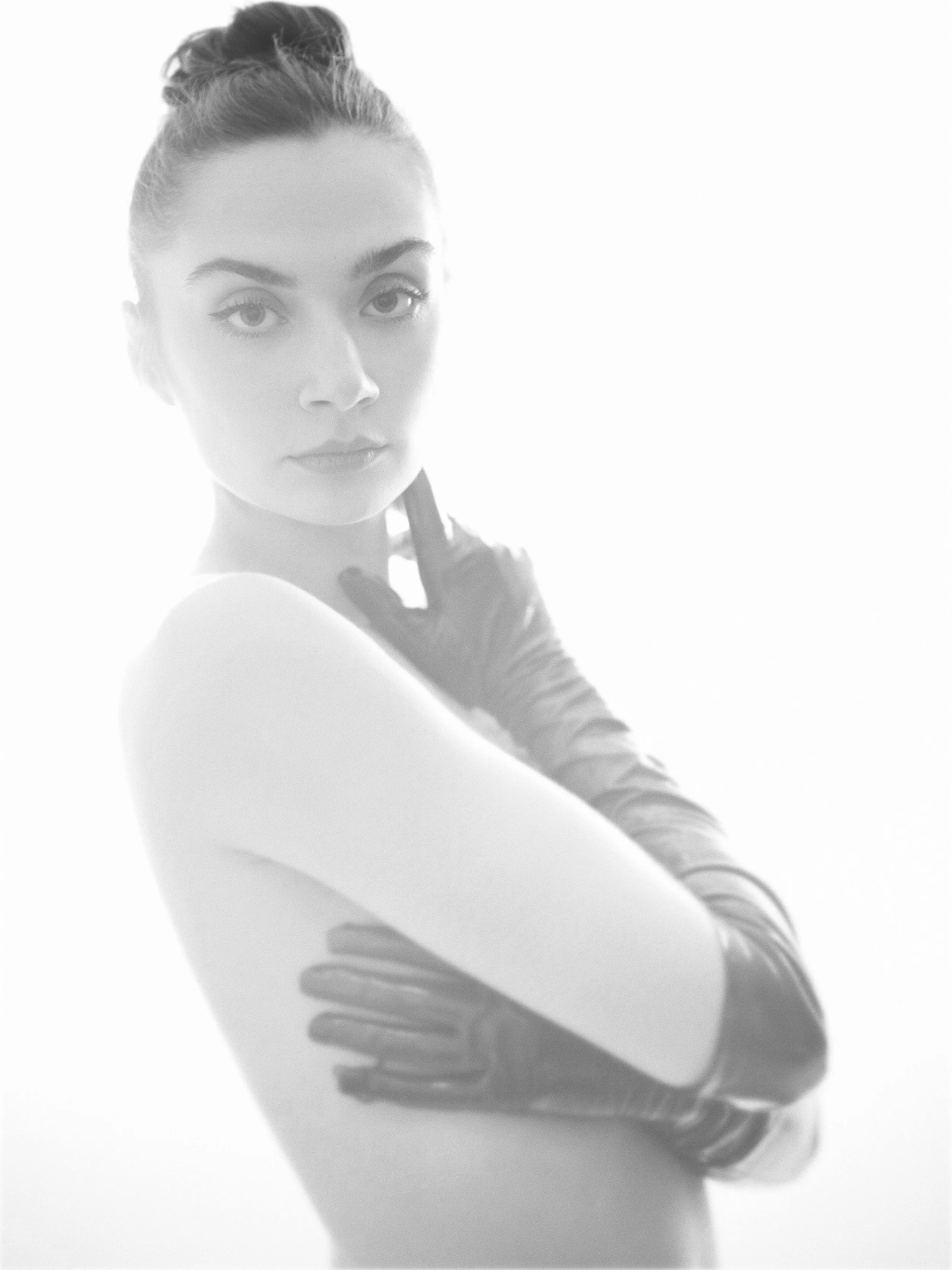 A woman with dark hair styled in a bun, wearing a sleeveless top and black gloves, looking directly at the camera with arms crossed.