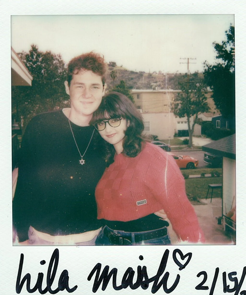 A young man and woman standing outdoors, smiling and posing for a photo. The man has curly hair, is wearing a black shirt with a star necklace, and the woman has dark hair, glasses, and is wearing a red sweater with a patch that says "RAGGED." In the