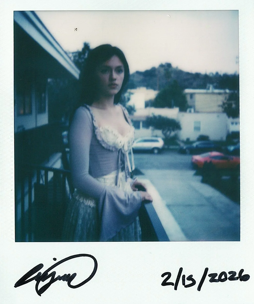A young woman in a vintage, historical-style dress with lace details, standing on a balcony with a railing, overlooking parked cars and buildings with hills in the background.