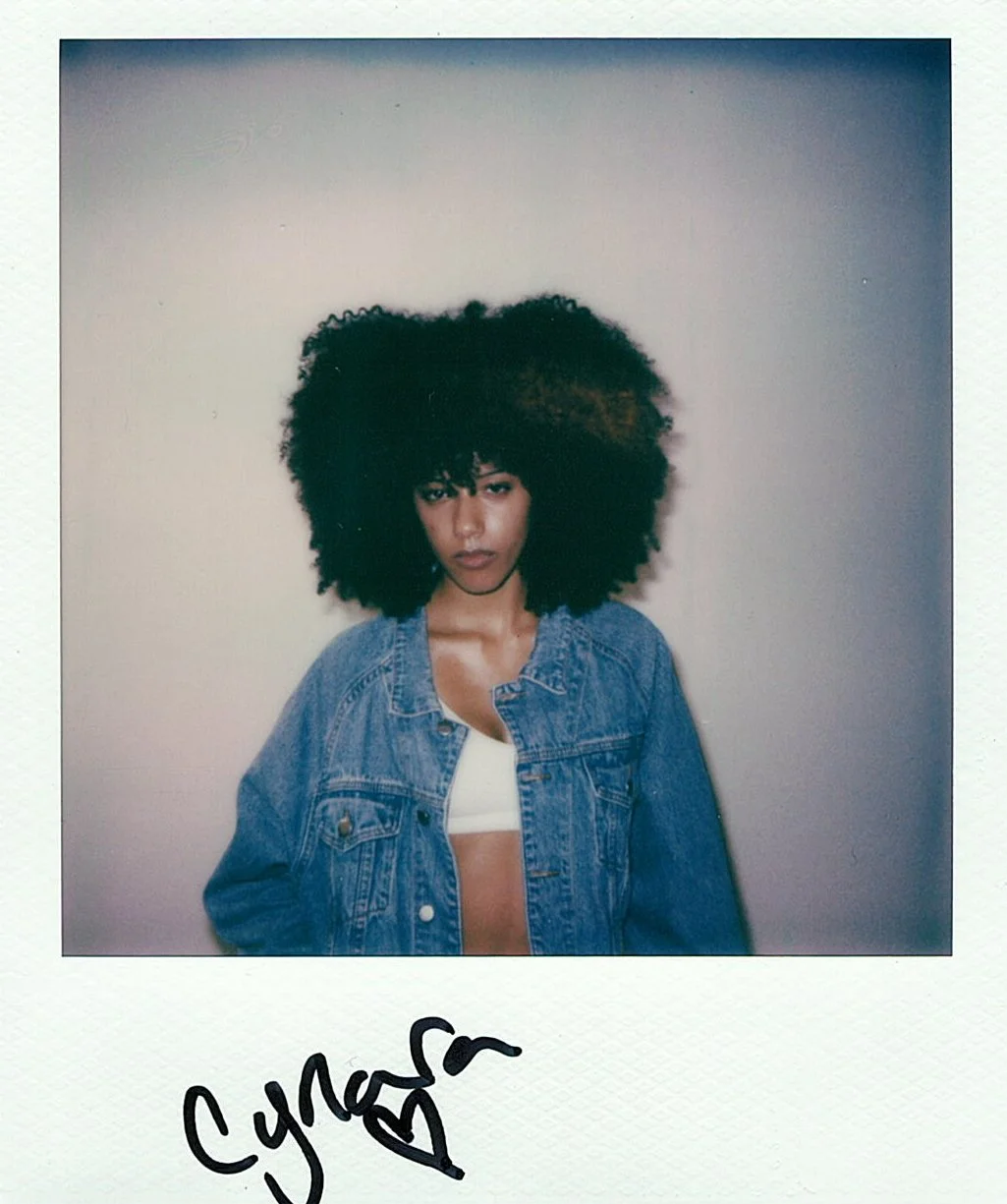 A woman with a large afro hairstyle standing against a plain wall, wearing a denim jacket over a white crop top.