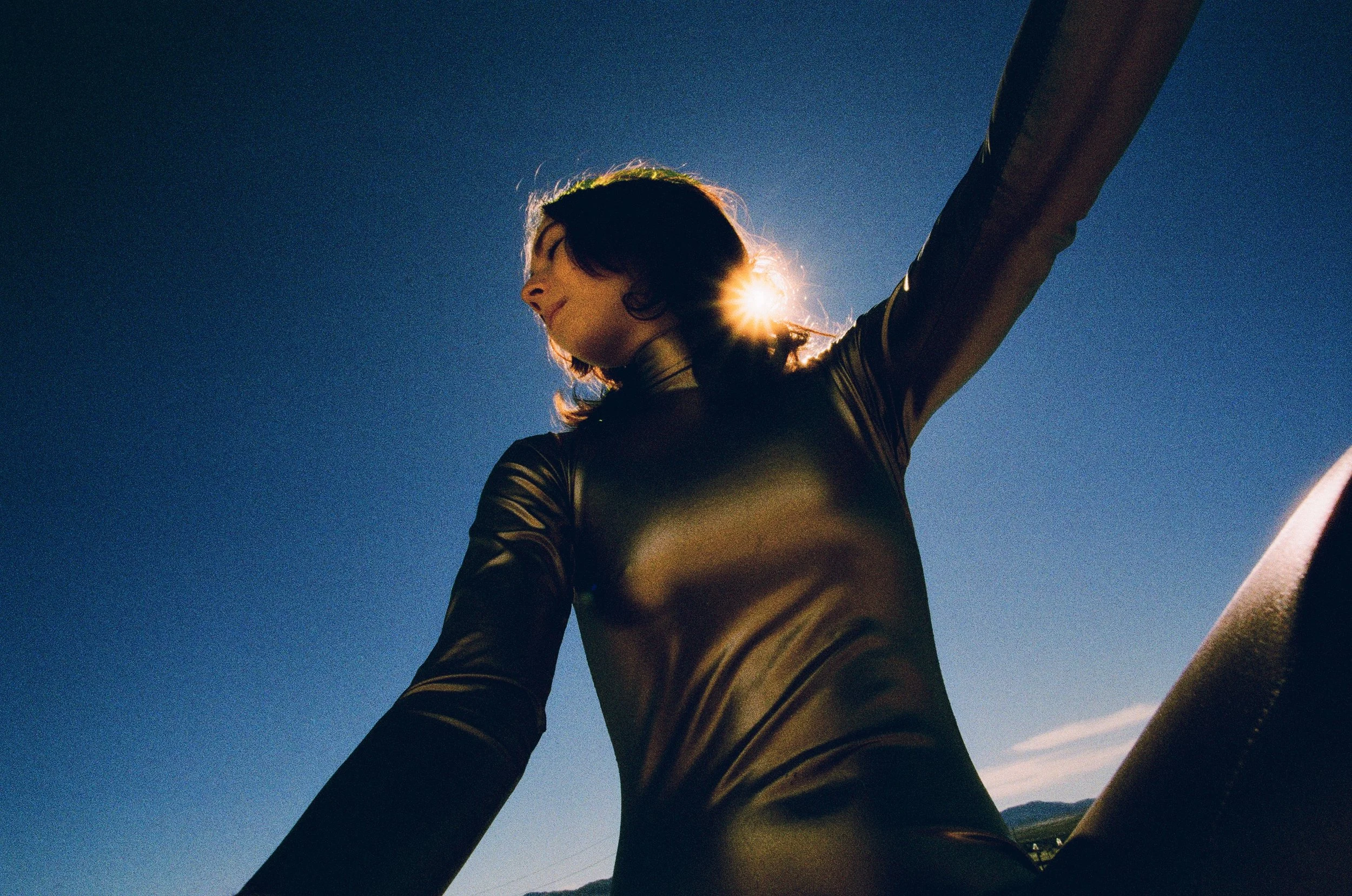 Woman wearing a shiny black outfit, sitting outdoors under a clear blue sky with the sun behind her head.