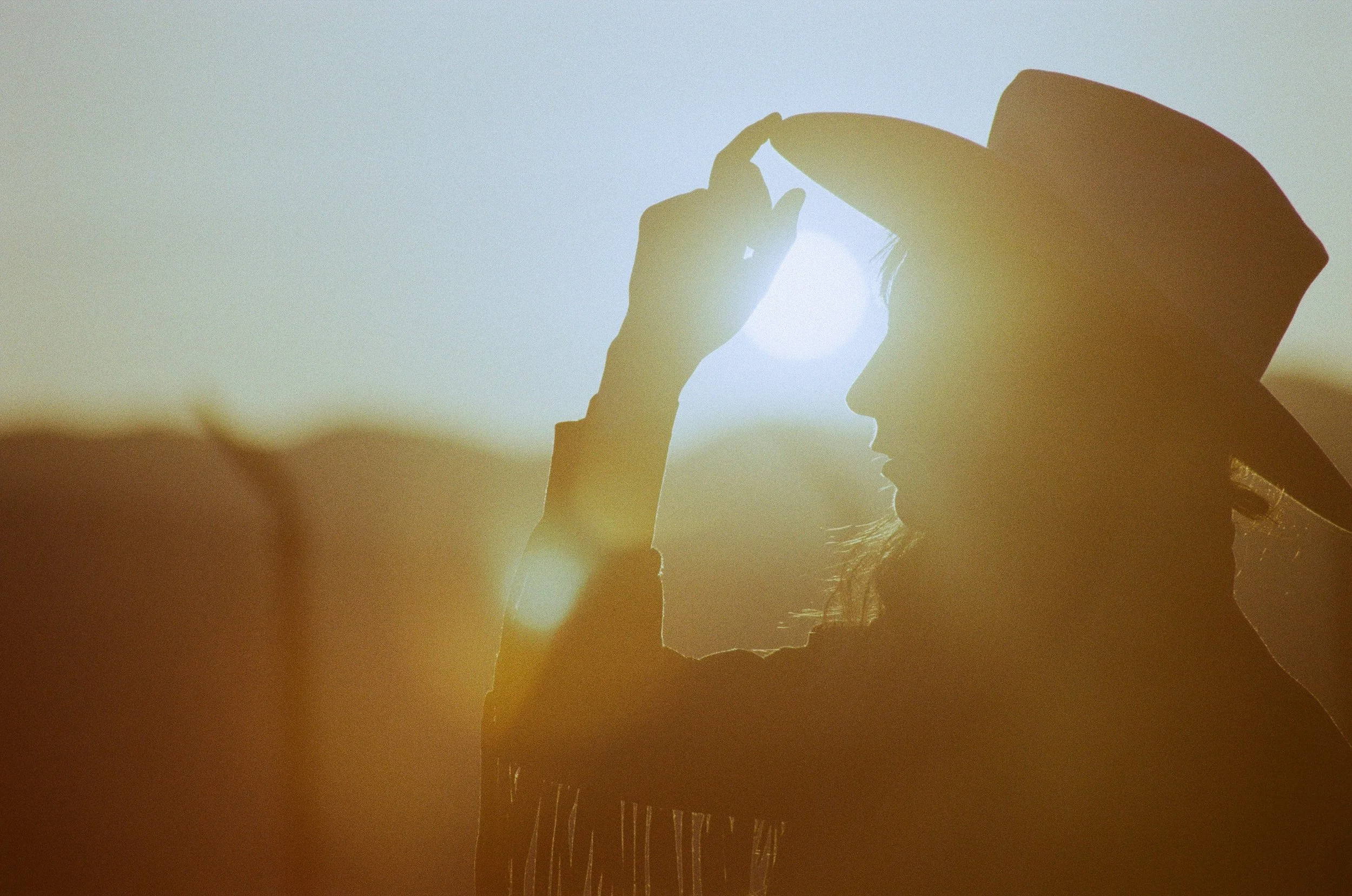 Silhouette of a woman wearing a wide-brimmed hat, adjusting it with her hand, against a bright sun in the sky.
