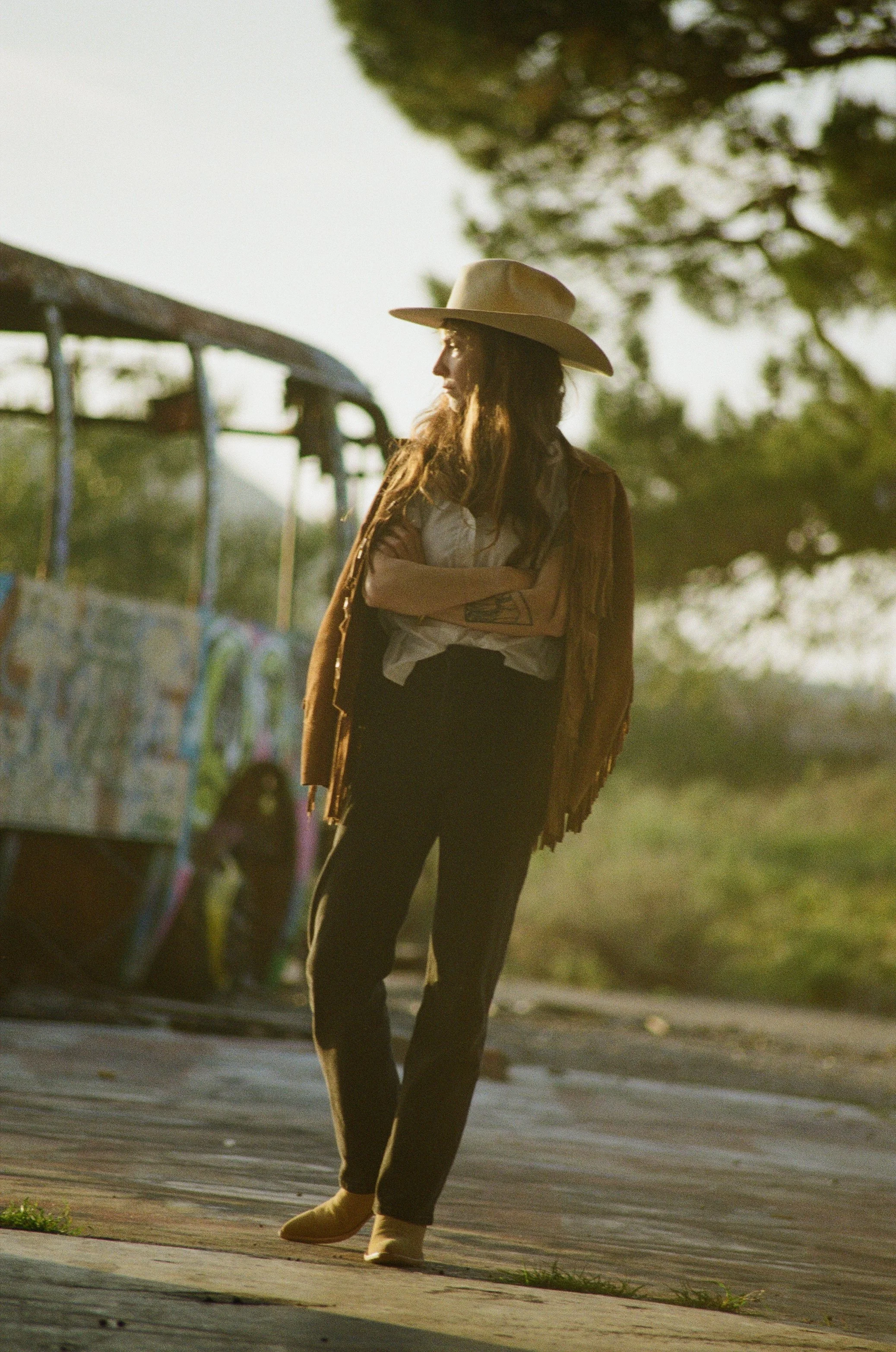 A woman standing outdoors in a cowboy hat, with her arms crossed, near a graffiti-covered vehicle, with trees and sunlight in the background.