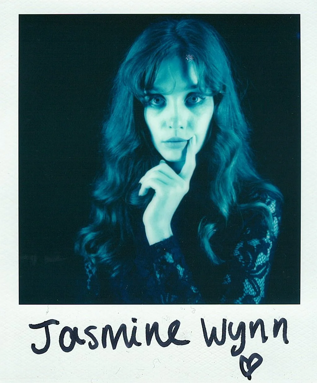 Polaroid photo of a woman with long wavy hair, posing with her finger on her lips, black lace top, against a dark background, with the name 'Jasmine Wynn' handwritten below and a small heart symbol.