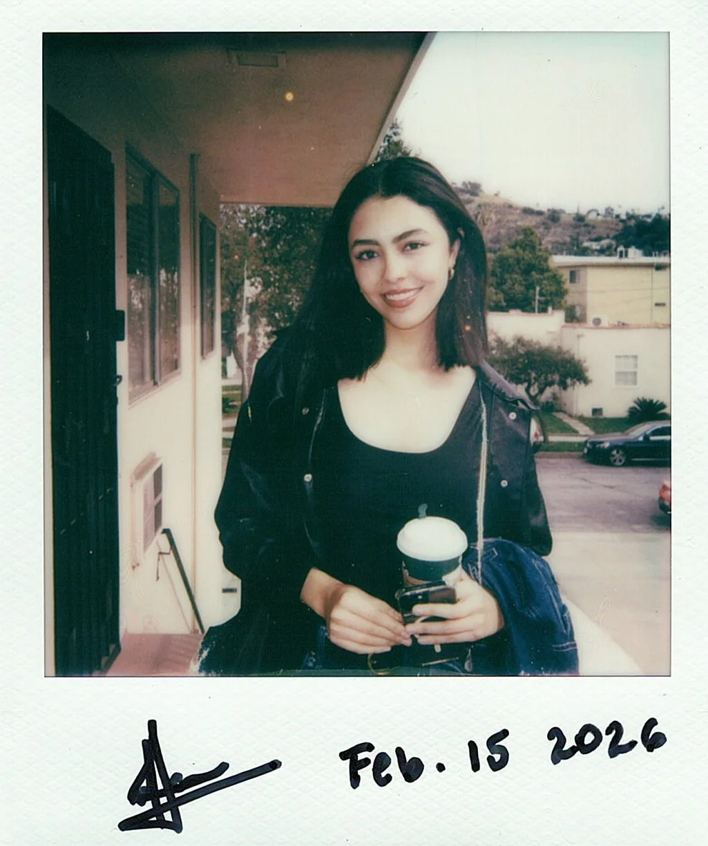A young woman with shoulder-length dark hair stands outdoors, holding a drink with whipped cream. She is smiling and wearing a black jacket over a black top, with a denim jacket draped over her arm. The background features a residential area with bui