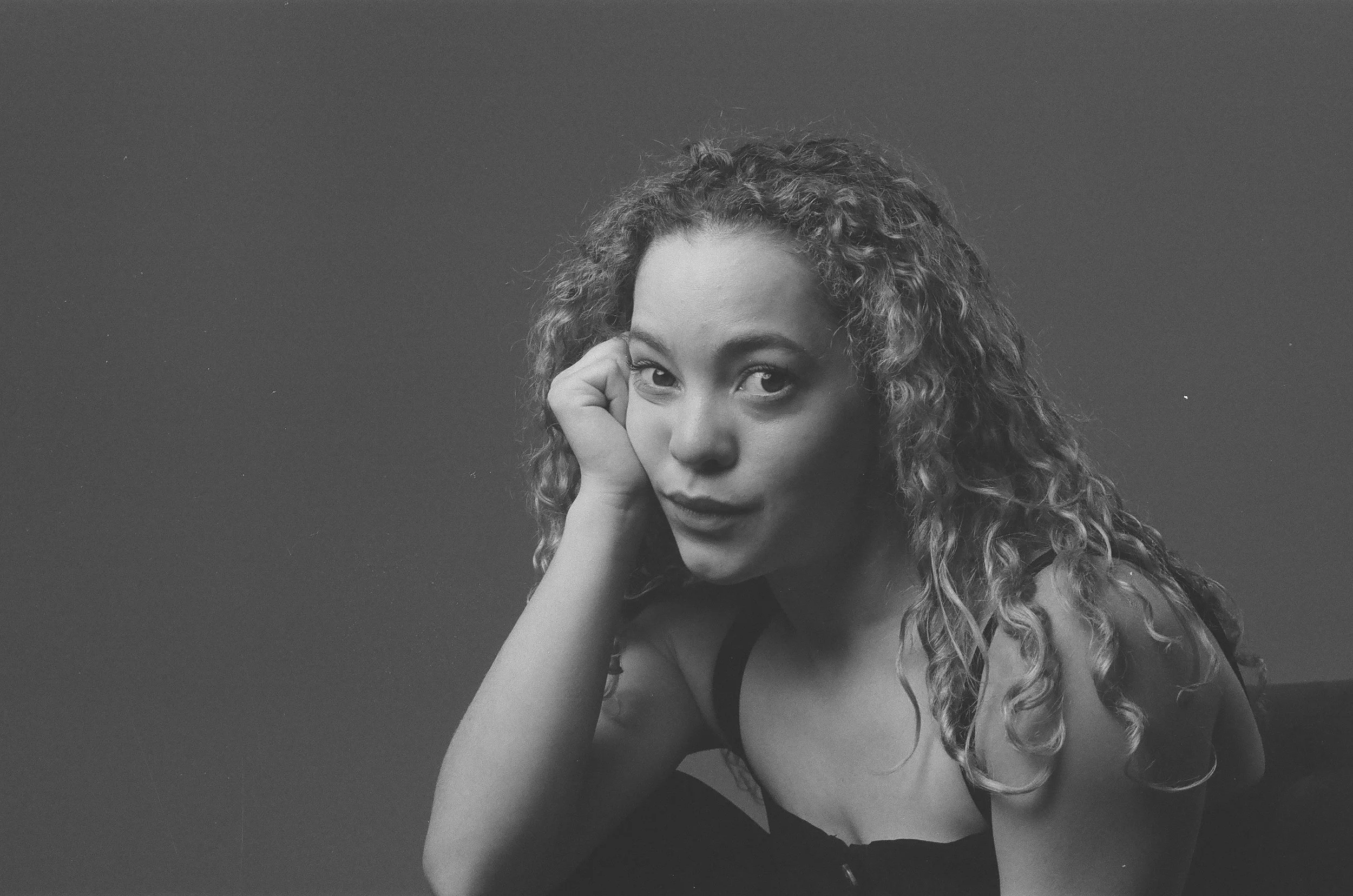 Black and white portrait of a young woman with curly hair, resting her face on her hand and looking at the camera.