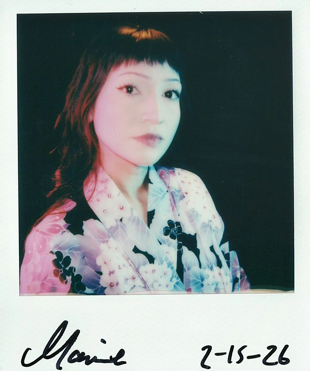 A woman with dark hair and bangs wearing a floral patterned shirt, posing against a dark background. The image is a color Polaroid with handwritten name 'Maisie' and date '2-15-26' at the bottom.