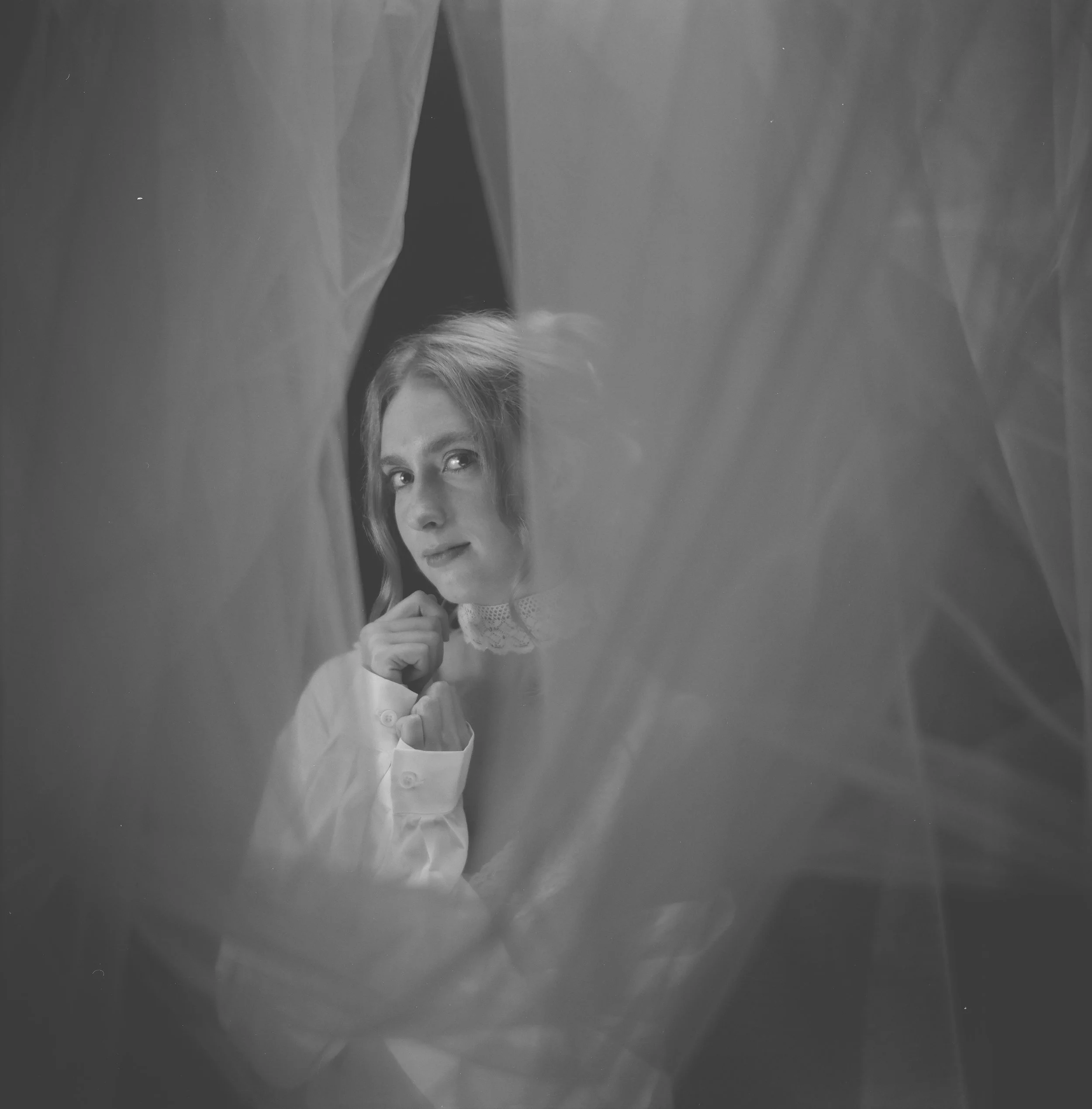 Black and white photo of a young woman with short hair, looking through a semi-transparent curtain or fabric, with her hand near her chin, wearing a white blouse with lace collar.