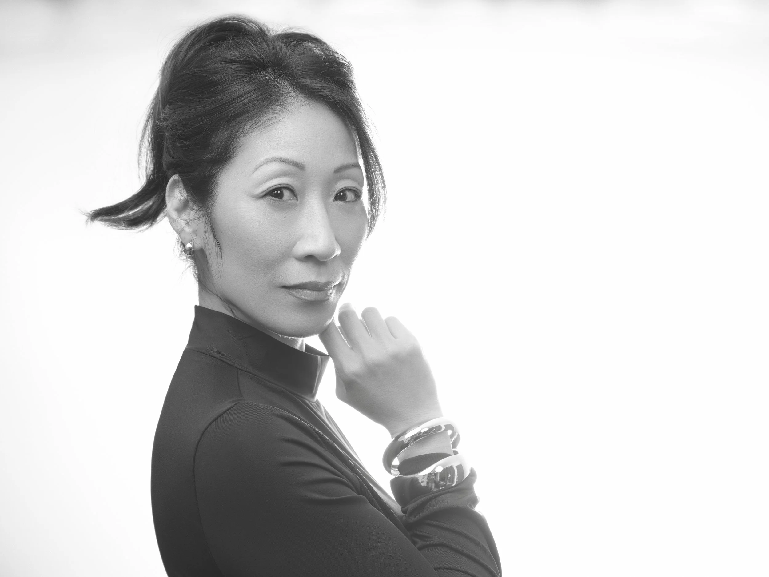 Black and white portrait of a woman with short hair, wearing a high-collared top and multiple bracelets, looking directly at the camera with her hand near her chin.