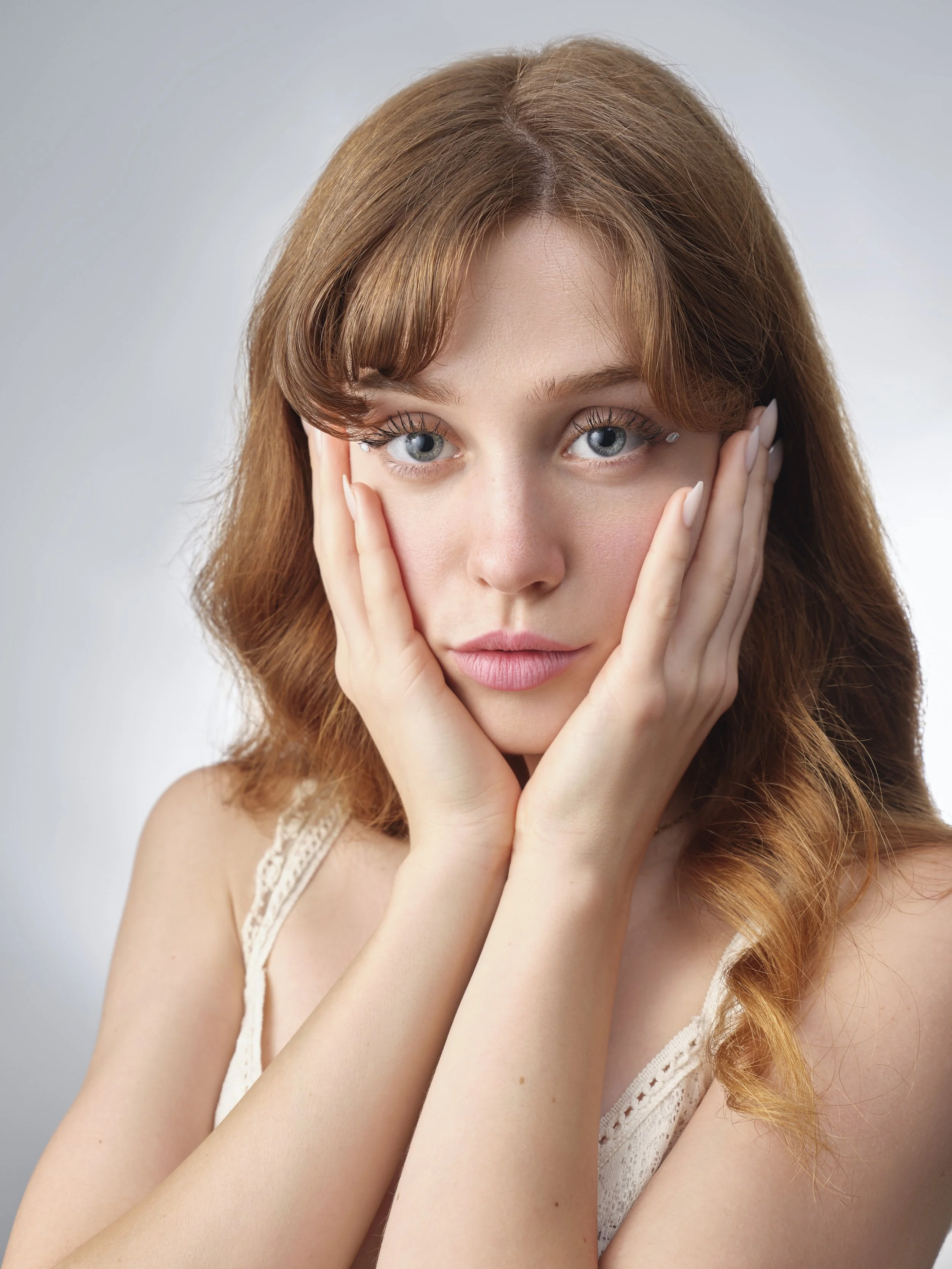 A young woman with red hair and blue eyes holding her face with both hands, looking directly at the camera.