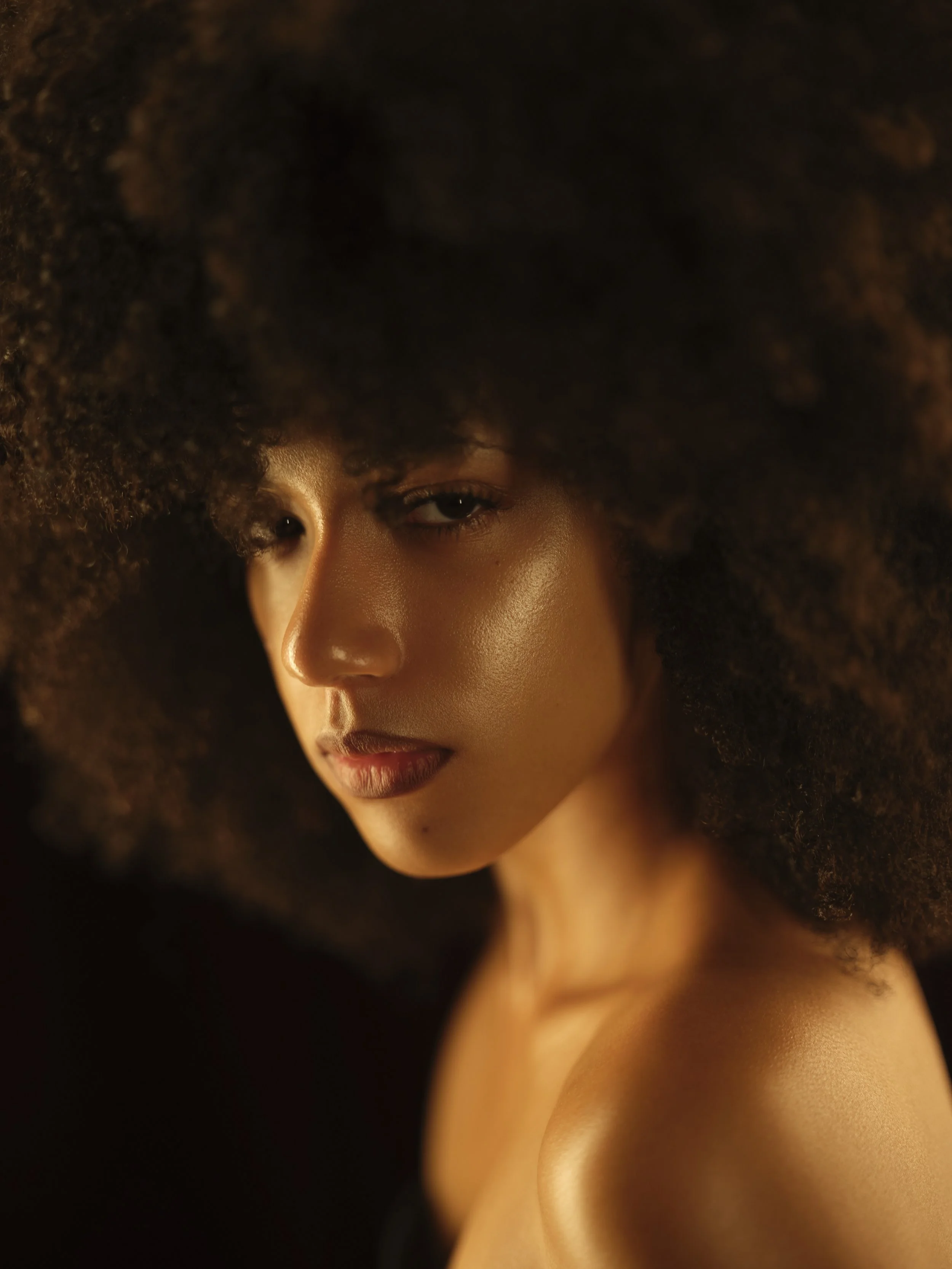 Close-up portrait of a woman with dark, curly hair, glowing skin, and a subtle smile, looking slightly downward.