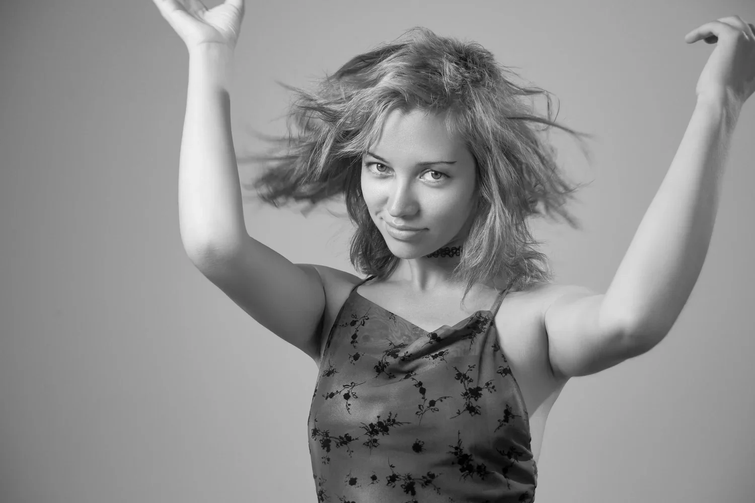 Black and white photo of a woman with tousled hair, wearing a floral dress and choker, looking confidently at the camera with arms raised.