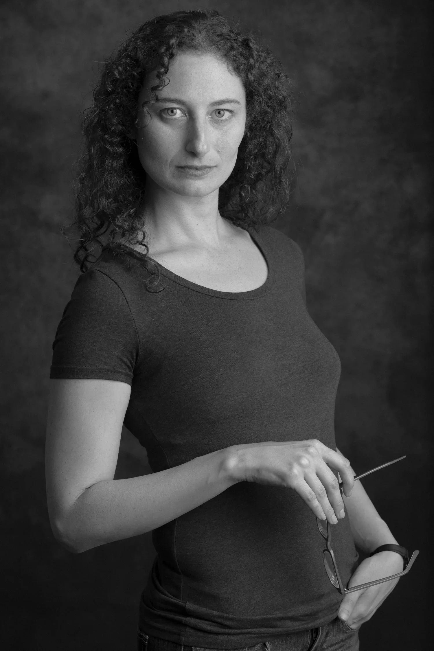 Black and white portrait of a woman with curly hair, wearing a dark t-shirt, holding glasses in her right hand, standing against a dark background.