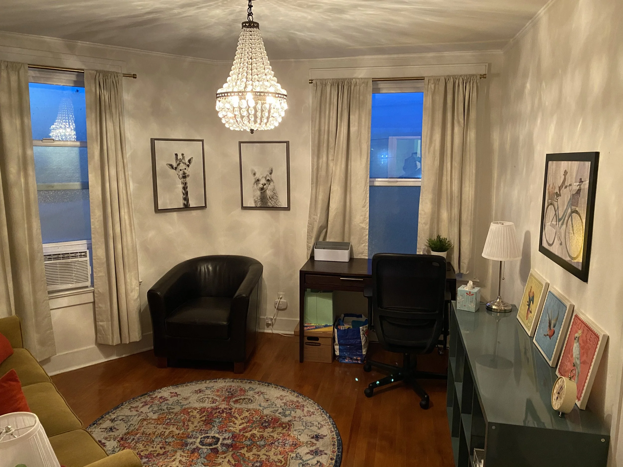 Living room with a chandelier, two windows with beige curtains, and various framed artworks including a giraffe and a bear on the wall. There is a black armchair, a yellow couch with orange pillows, a dark wooden desk with an office chair, a green sh