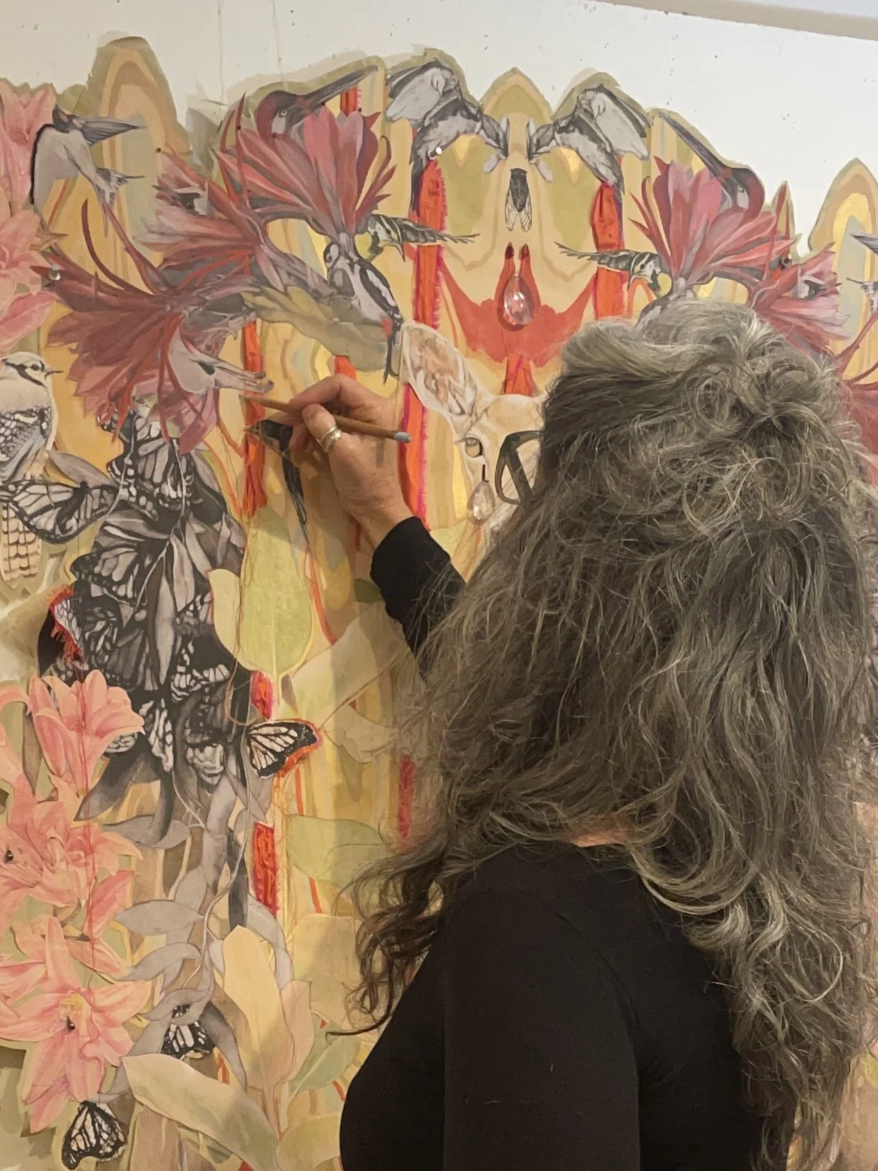 The artist working on adding details to intricate collage art piece featuring hummingbirds, butterflies, flowers, and other natural elements.