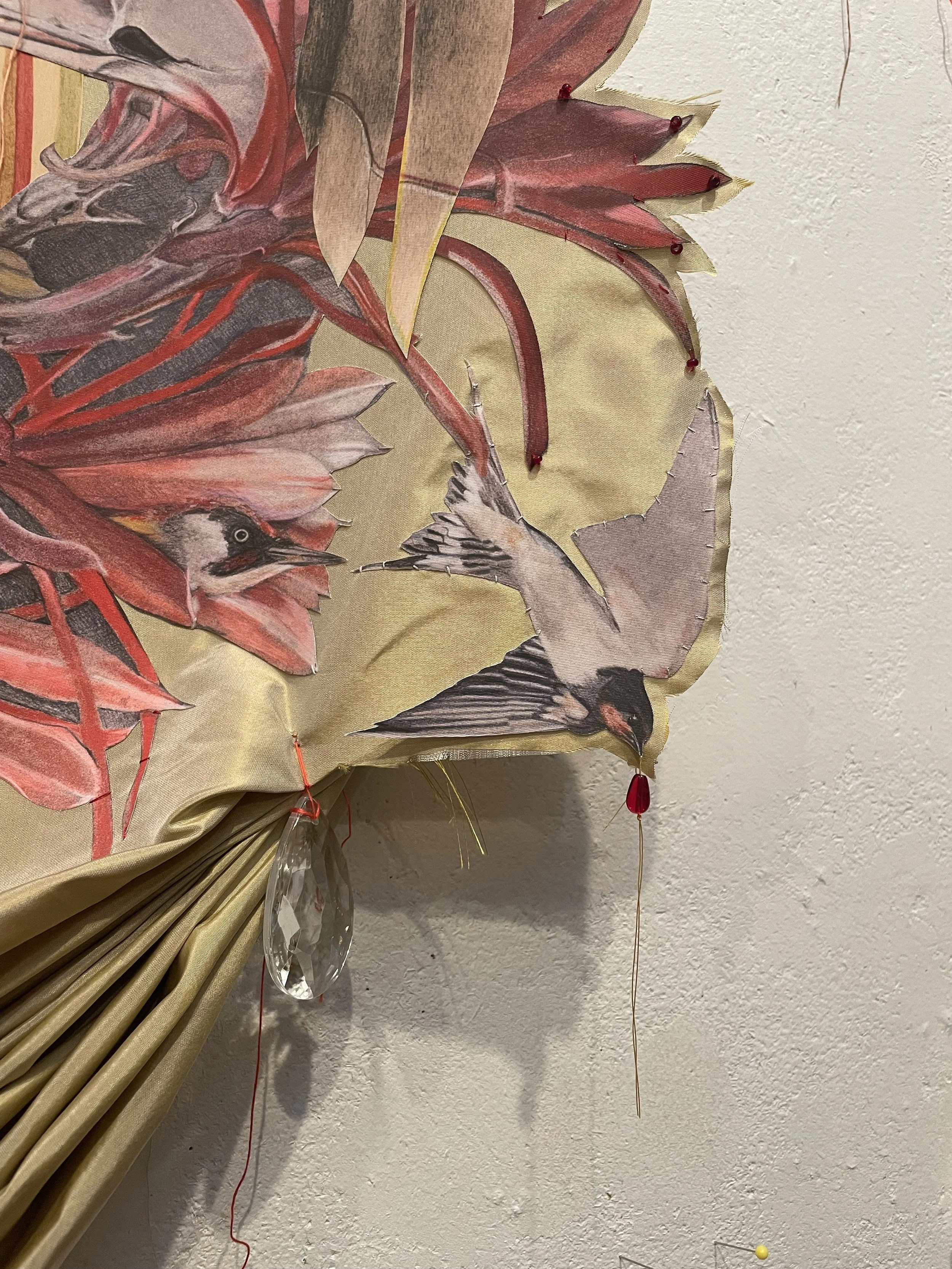 Detail featuring birds and flowers accented by red beads and dangling yellow and red threads, hanging on a plain white textured wall.