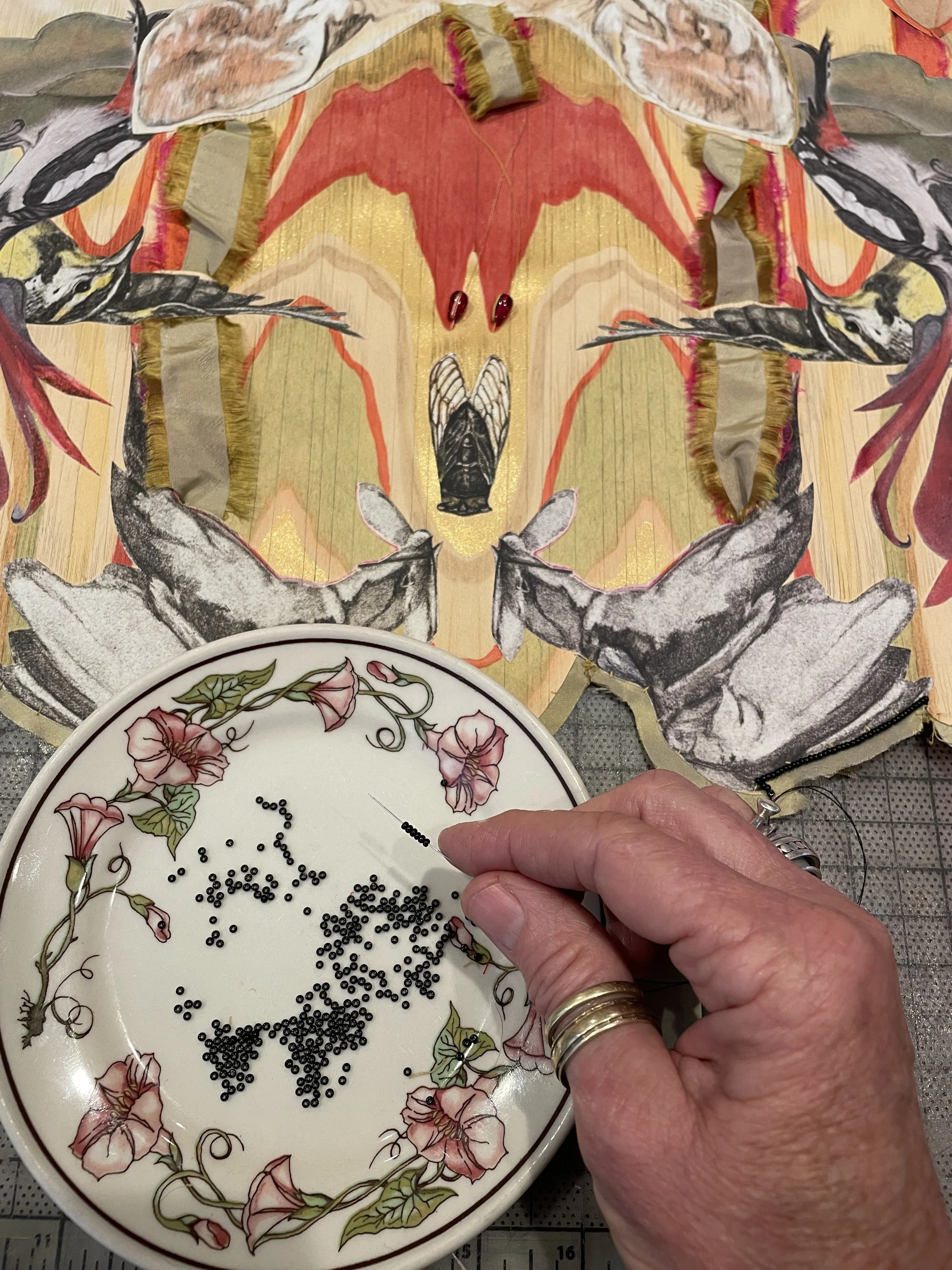 The artist's hand holding a needle and black seed beads above a small decorative ceramic dish with pink flowers and green leaves, on a table. 