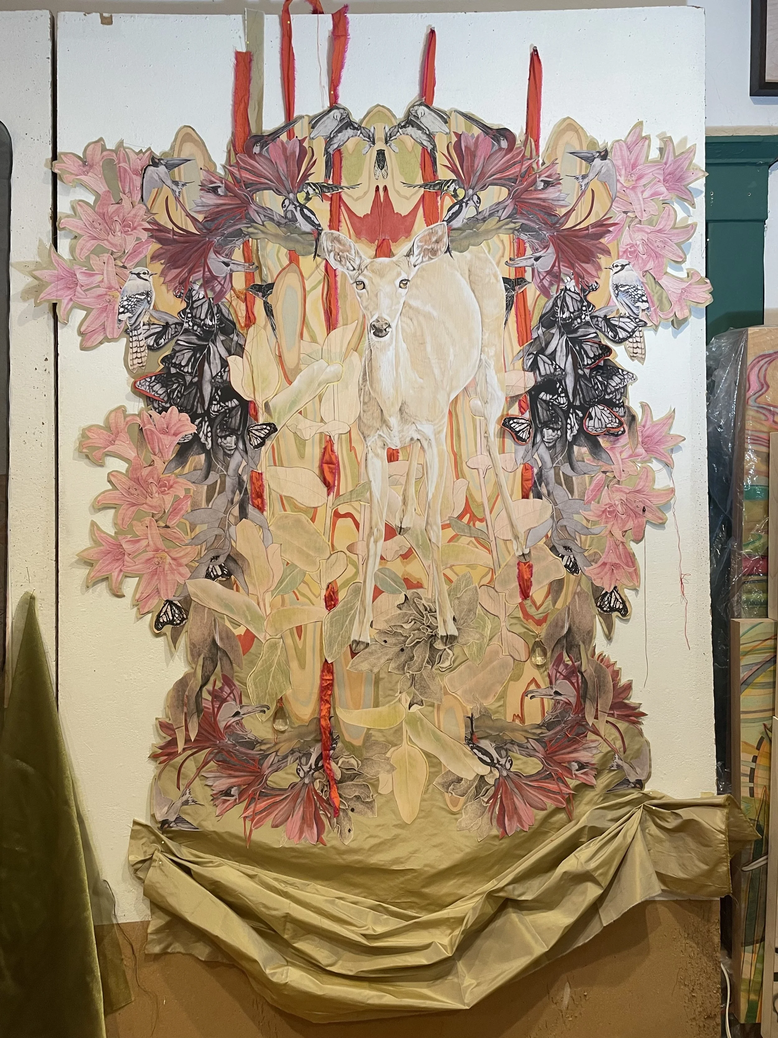 Work in progress on "The Return", a fabric collage with a central image of a white tailed deer surrounded by pink lilies, hummingbirds, and butterflies, with a symmetrical design, displayed on a studio wall.