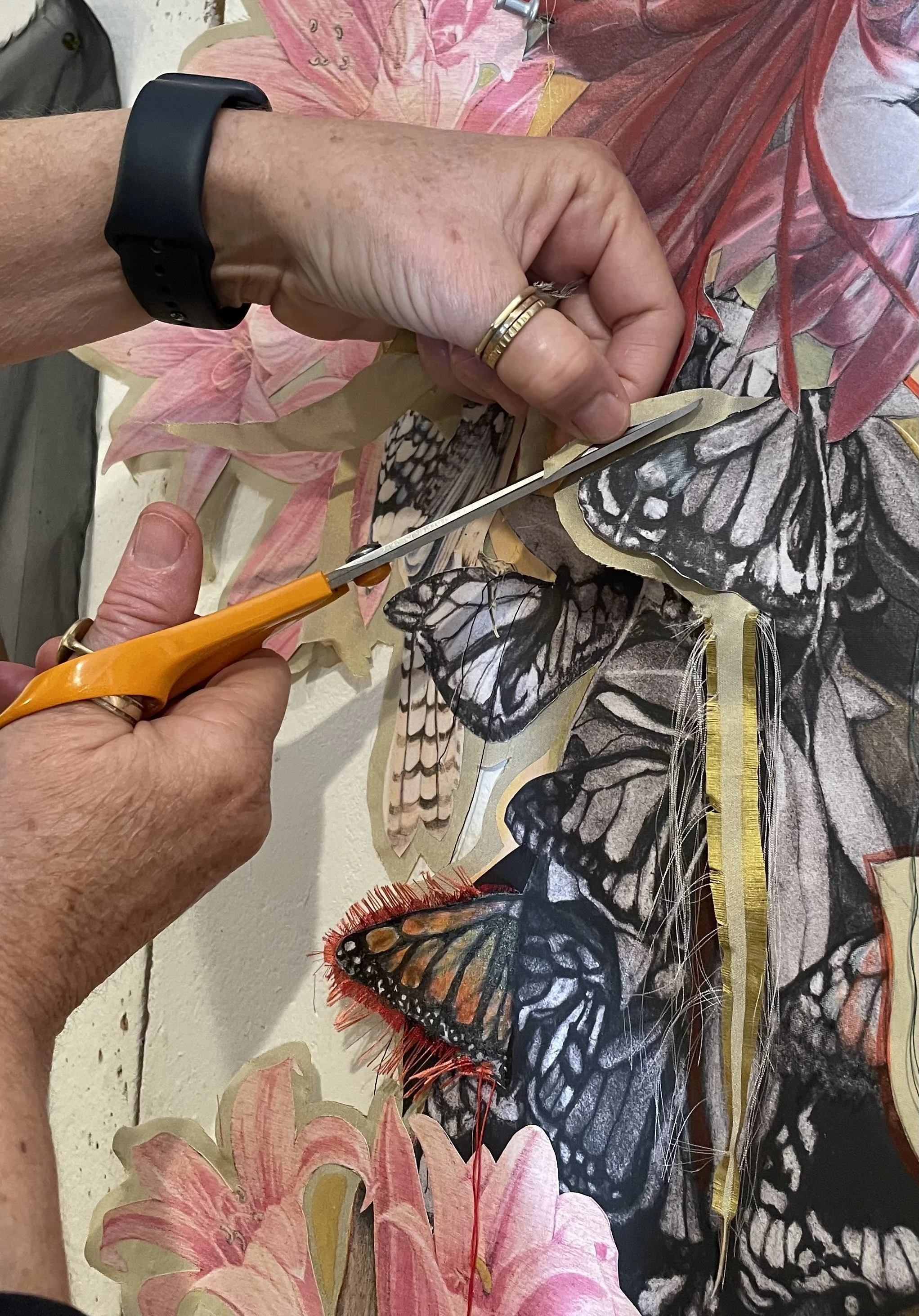 Detail view of the artist cutting out fabric with scissors surrounding butterfly and floral imagery in the background.