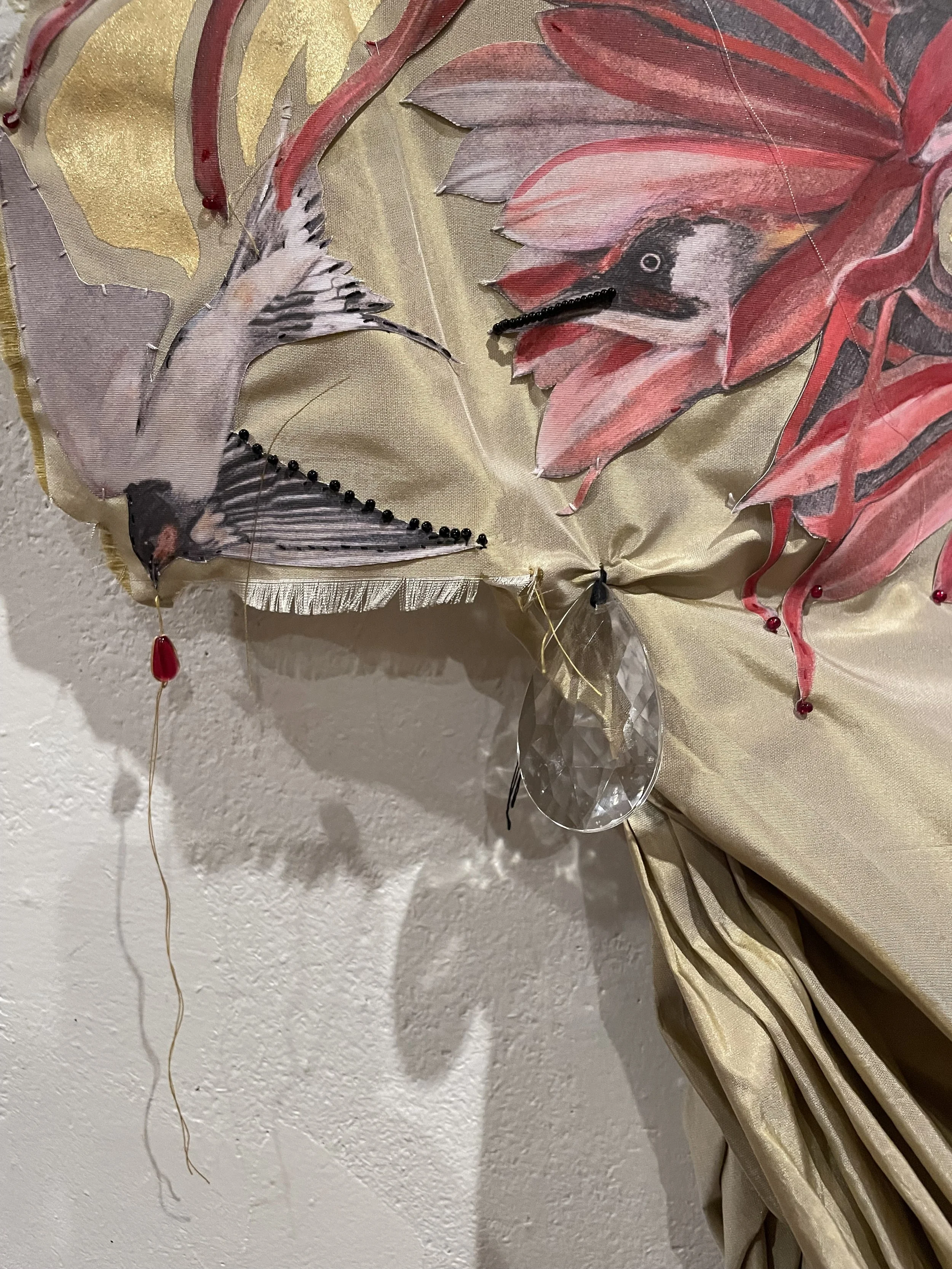 Detail of "The Return" featuring two birds, one with a long beak and black markings on white feathers, and the other with a prominent red and black head, surrounded by red and gold leaves, with some beads and a clear glass teardrop shaped beads.