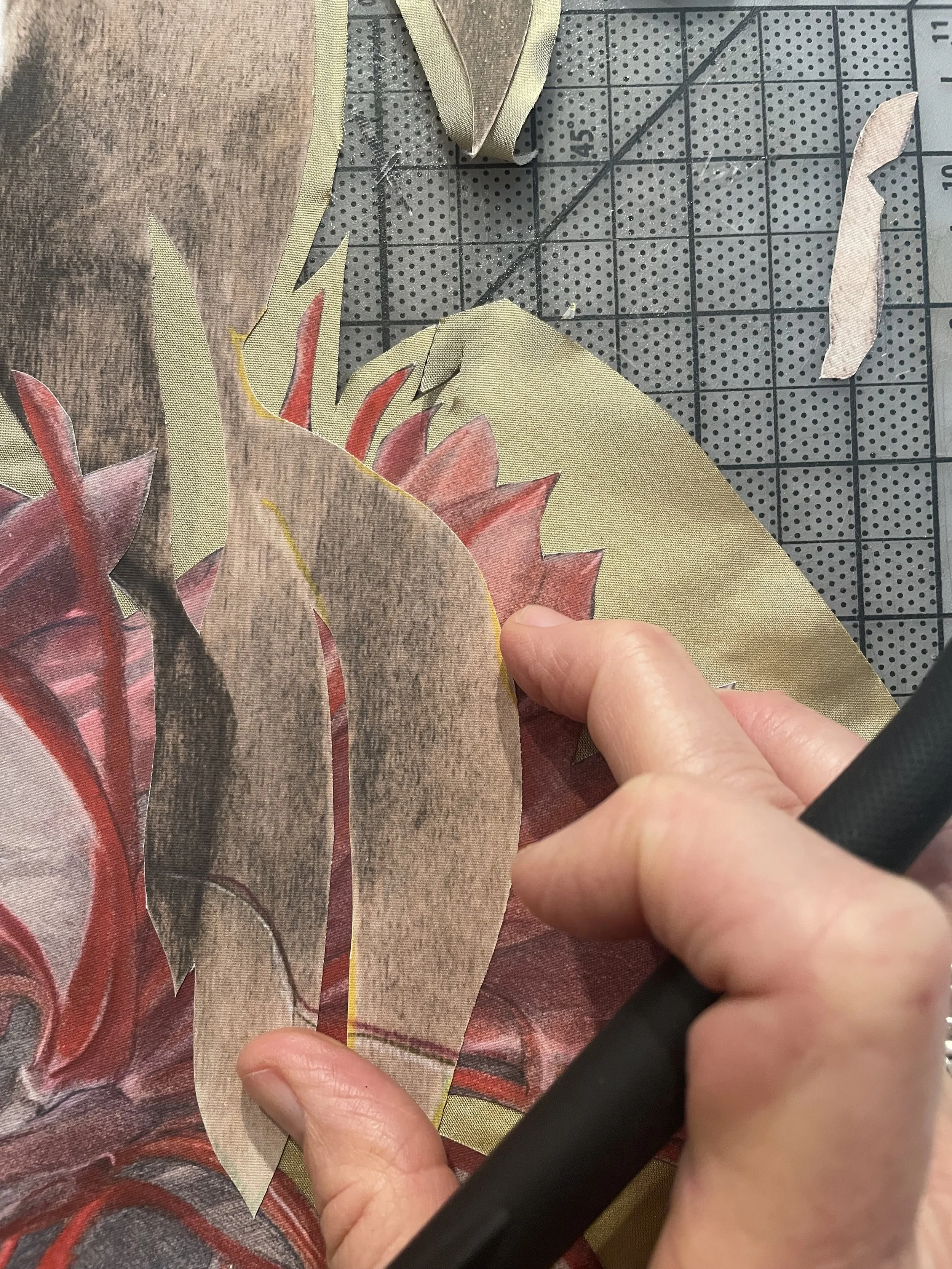 Close-up of the artist's hand holding an X-acto knife over cut-out fabric with petal-like patterns, placed on a grey cutting mat with measurement markings.
