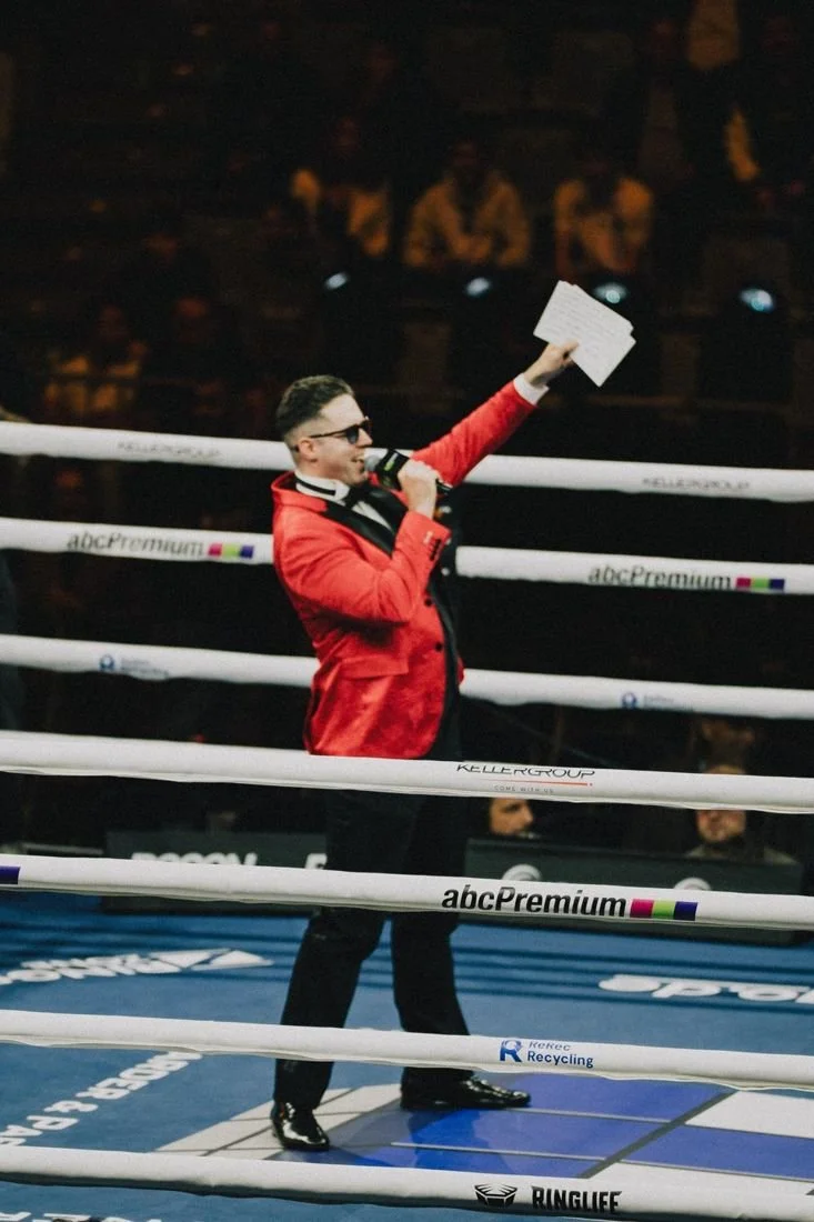 Tobias Greenhalgh in a red jacket in the boxing ring, announcing with microphone and cards raised