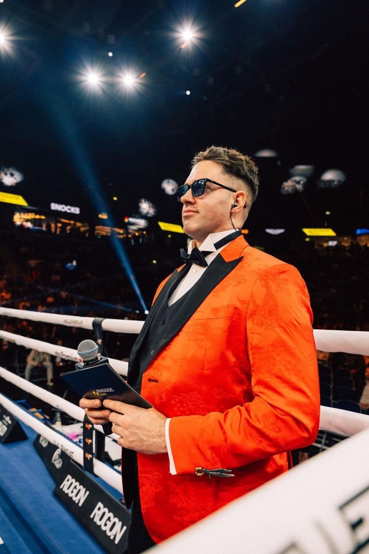 Tobias Greenhalgh ringside in an orange jacket and bow tie, holding a microphone and fight card
