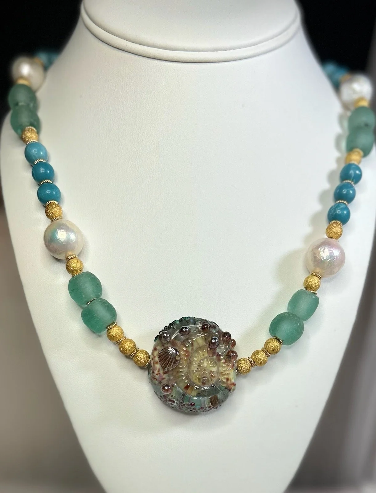  Handmade glass “shell” with green African handmade glass beads, six high quality 15 mm white freshwater pearls, blue Caribbean Larimor beads and golden dusted beads.  24” in length. 