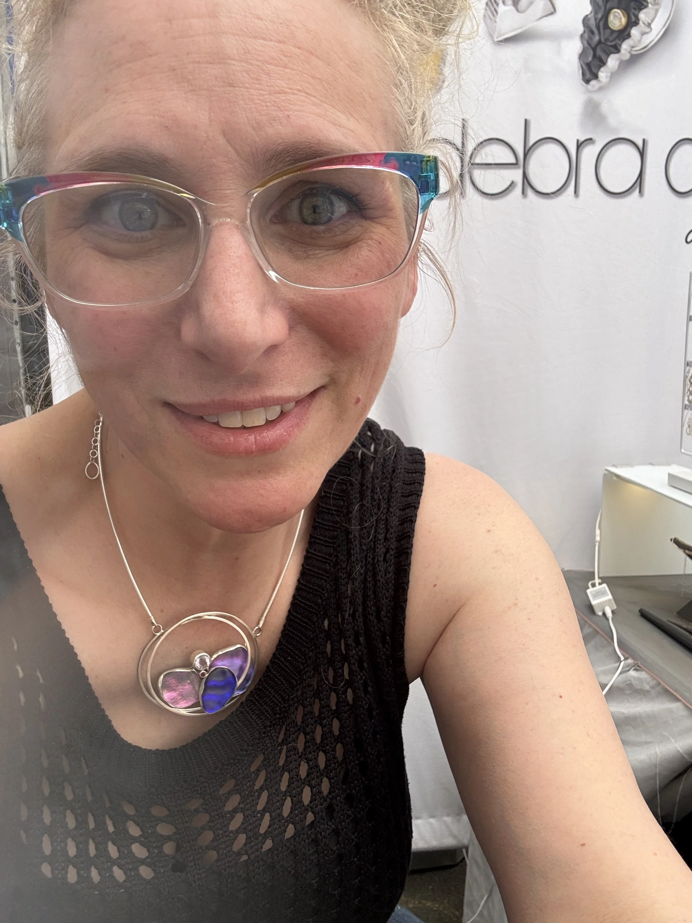 Artist, Debra Adelson in her jewelry booth wearing rainbow glasses.