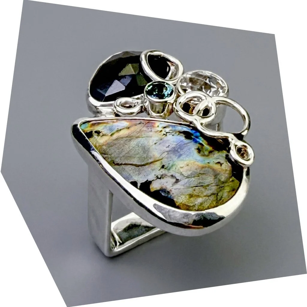 A silver ring with multiple gemstones, including a large iridescent shell, black, clear, and green stones, and silver loops, on a gray background.
