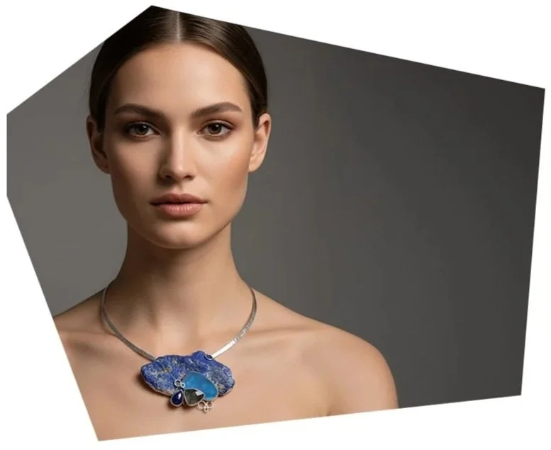 A woman with light skin and dark hair wearing a statement necklace with a large blue and purple pendant, posing against a dark gray background.