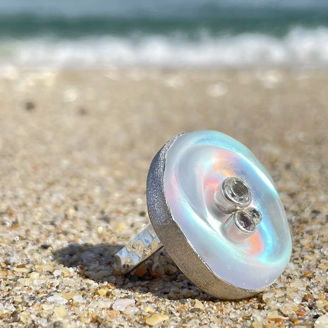 An image of a carved glass, sterling silver, and topaz ring on the beach with the caption, "Ocean inspired, not ocean made".