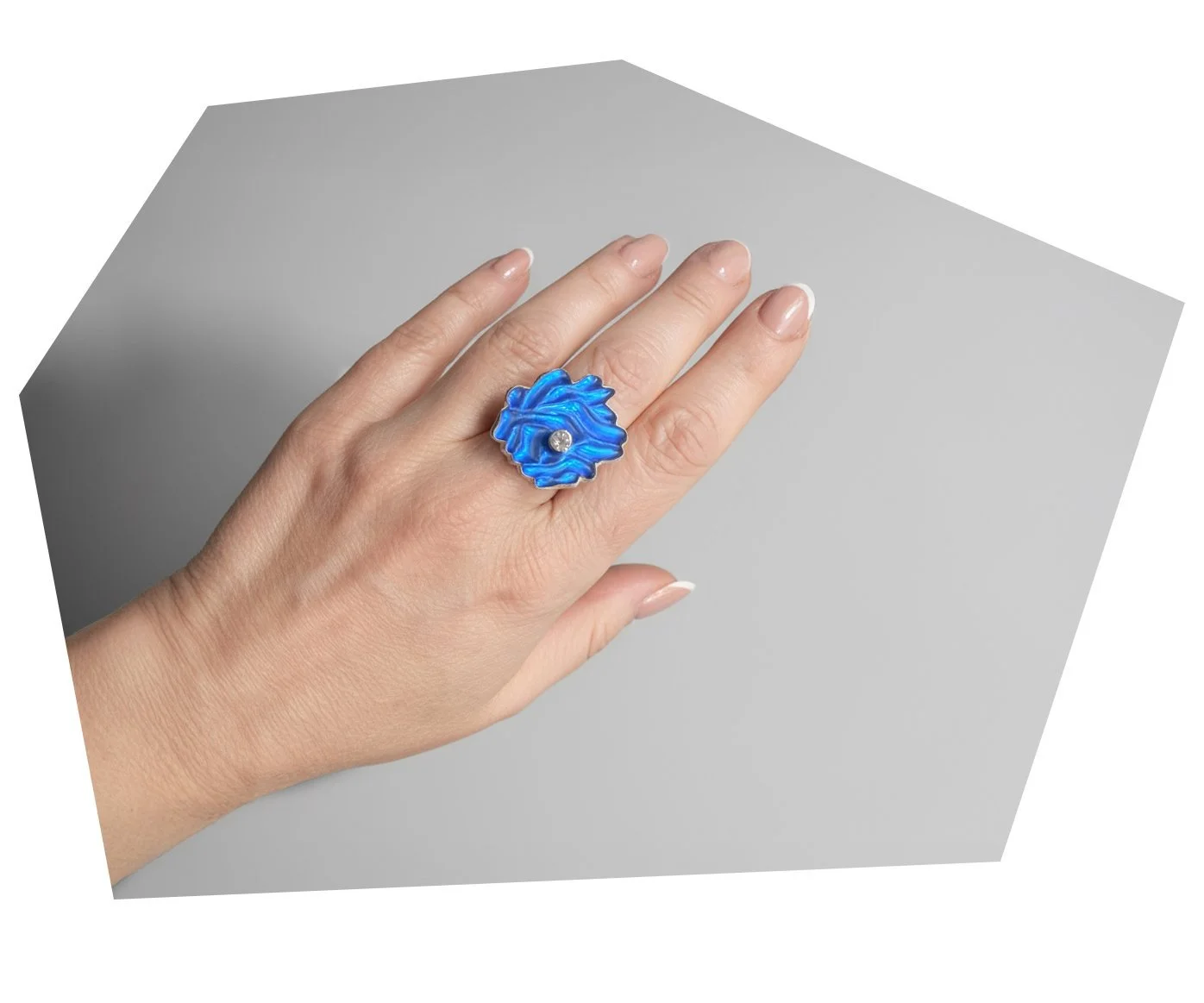 Close-up of a hand wearing a large blue floral-shaped ring with a small clear gemstone in the center, against a plain gray background.