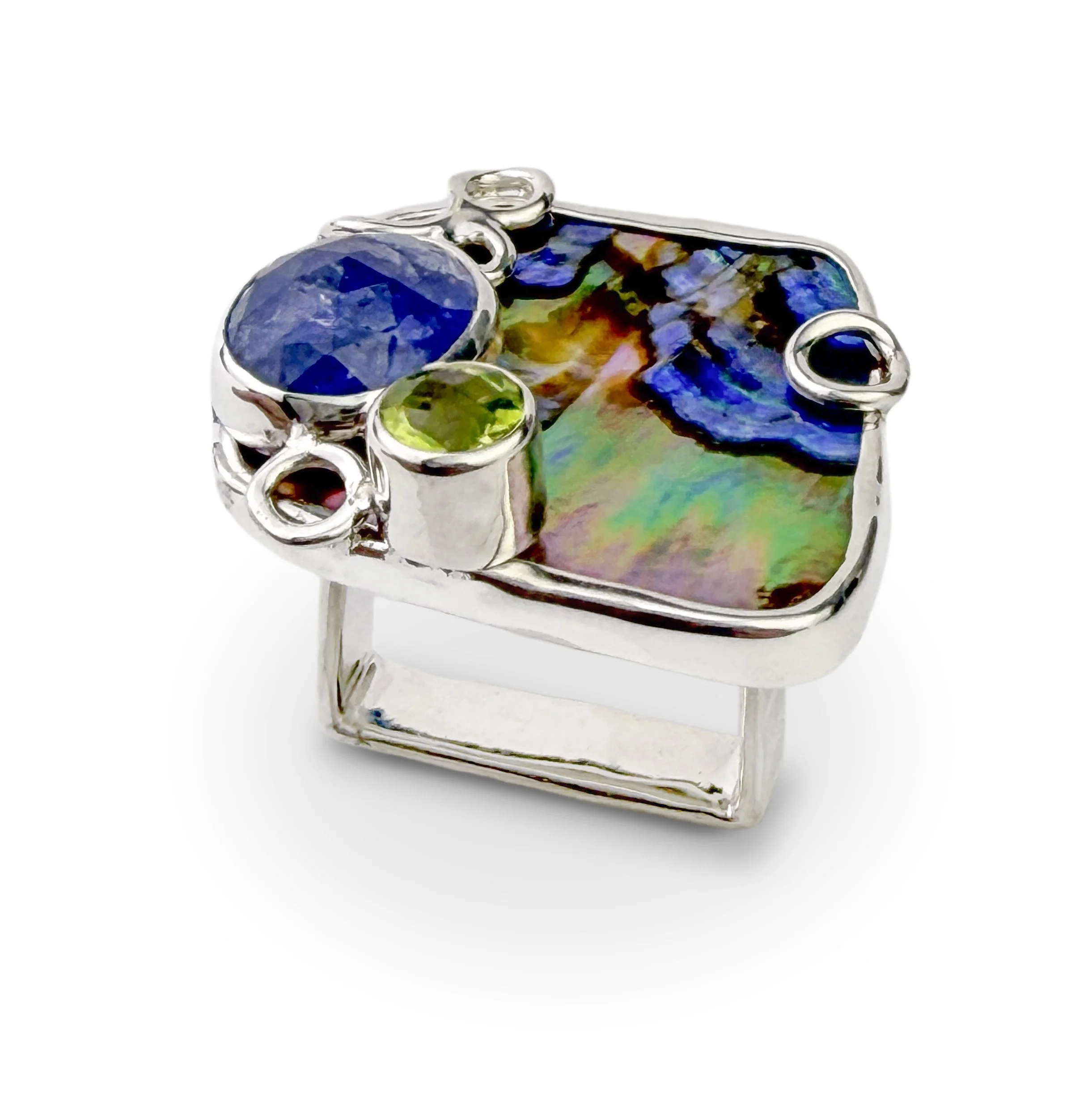 Front view of a silver mermaid statement ring featuring a colorful iridescent abalone shell backdrop with a faceted blue tanzanite and bright green peridot.