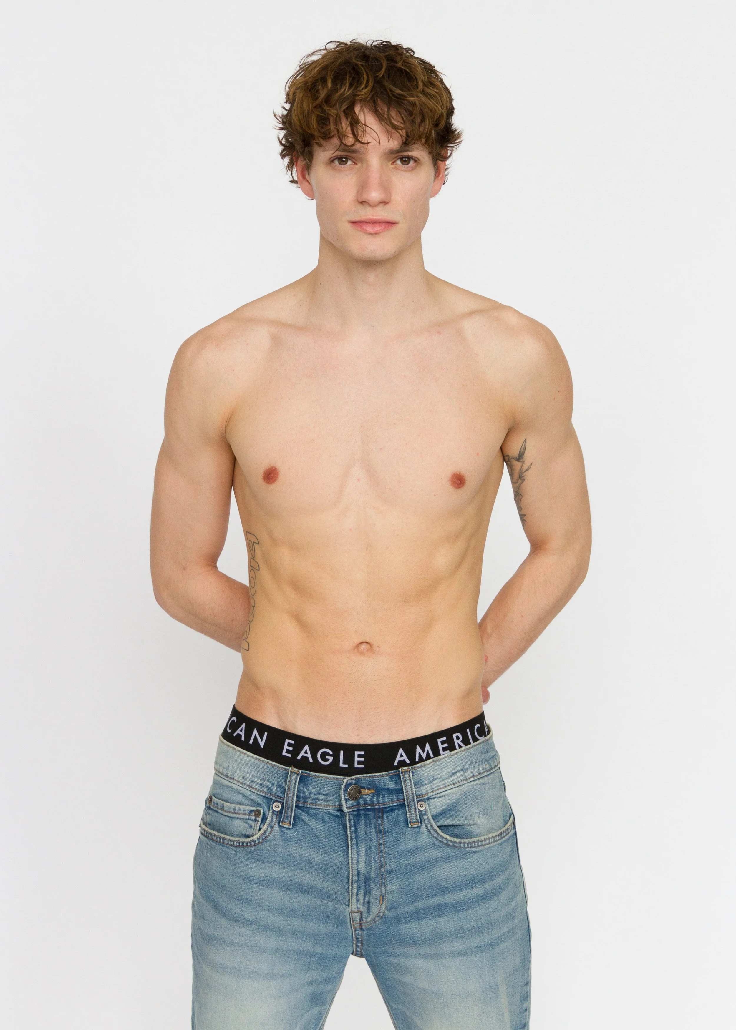A shirtless young man with brown curly hair, standing with hands behind back, wearing jeans and a waistband with 'American Eagle' logo, and has tattoos on arms, looking at the camera against a white background. Model Digitals
