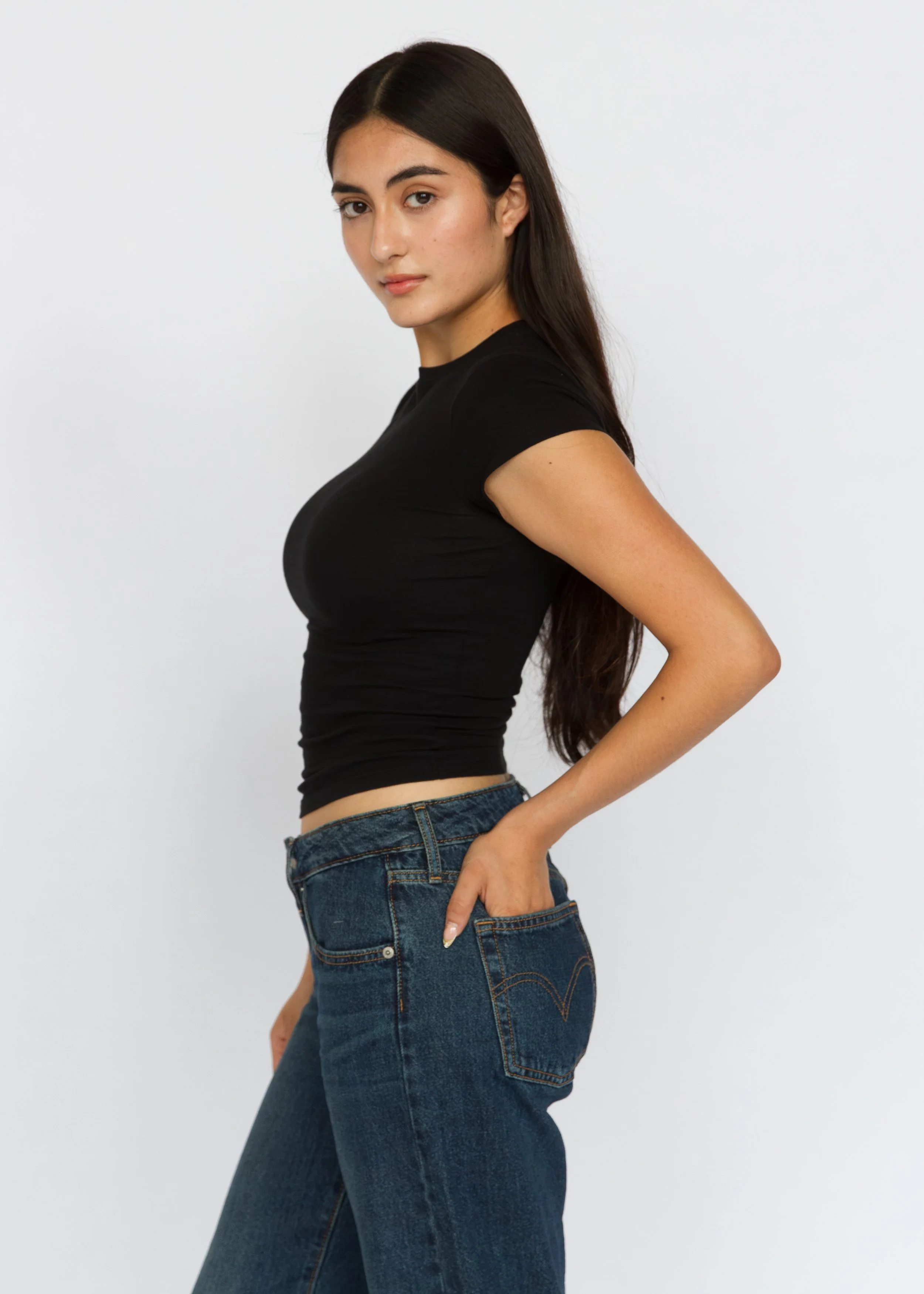 A woman with long dark hair wearing a black t-shirt and blue jeans standing against a plain light background, posing with one hand on her hip. Model Digitals