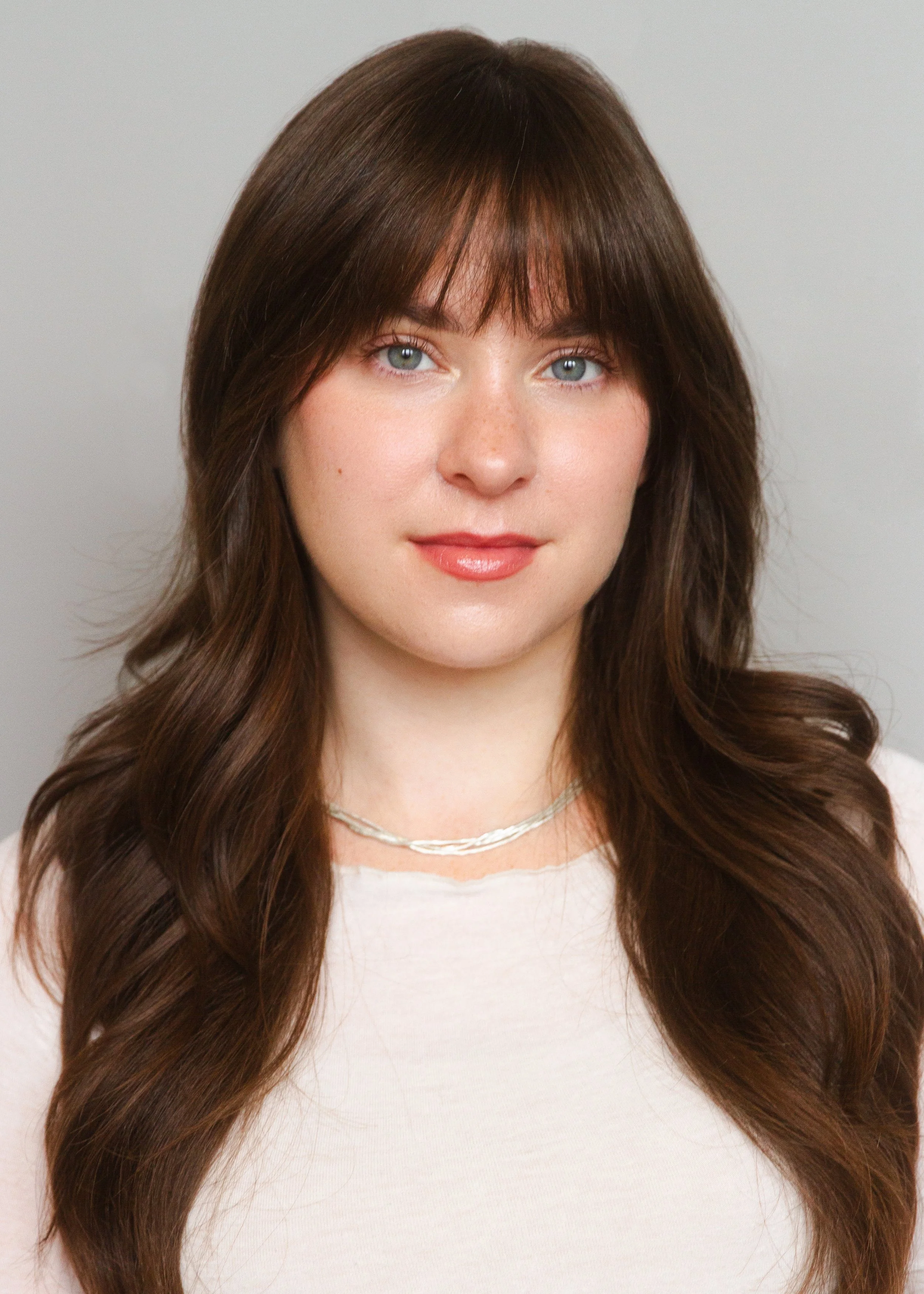 A young woman with long, wavy brown hair, light blue eyes, and fair skin, wearing a white top and a silver necklace, posing against a neutral gray background.