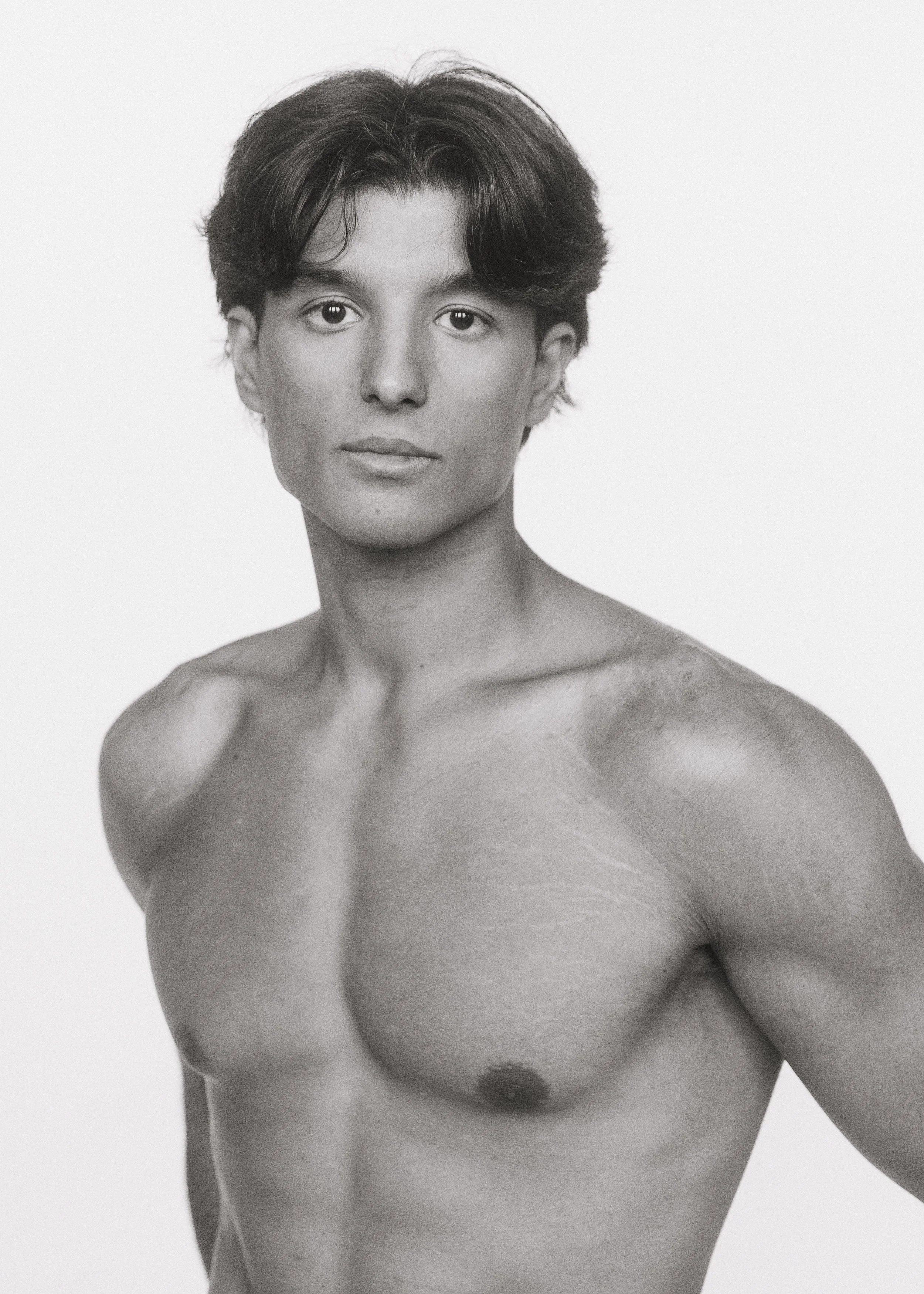 Black and white portrait of a young man with short, wavy hair, looking directly at the camera with a neutral expression, shirtless, showcasing a muscular chest and shoulders against a plain white background.