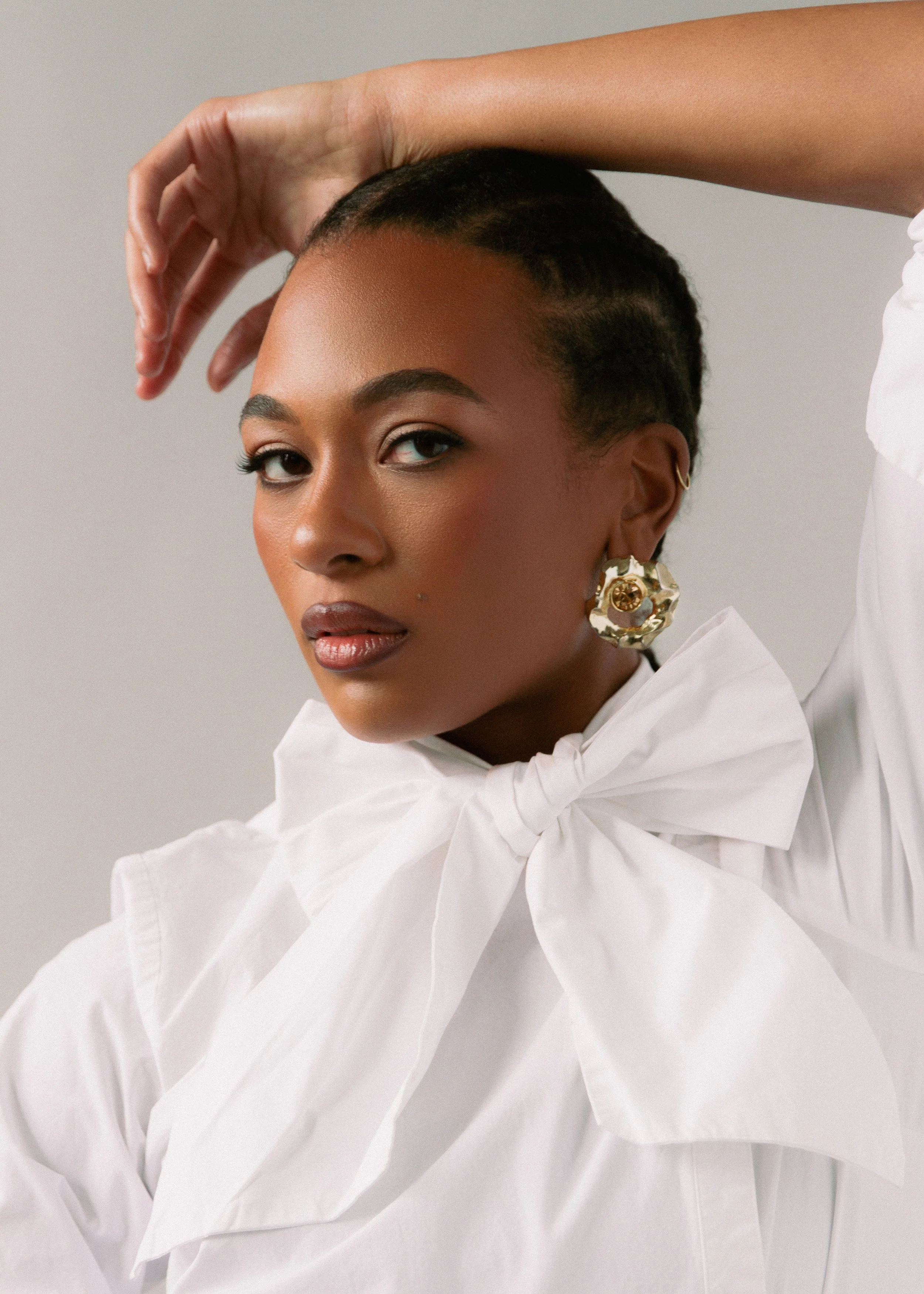 A woman with dark hair styled back, wearing gold hoop earrings and a large white bow tie blouse, posing with her arm raised behind her head against a light grey background.